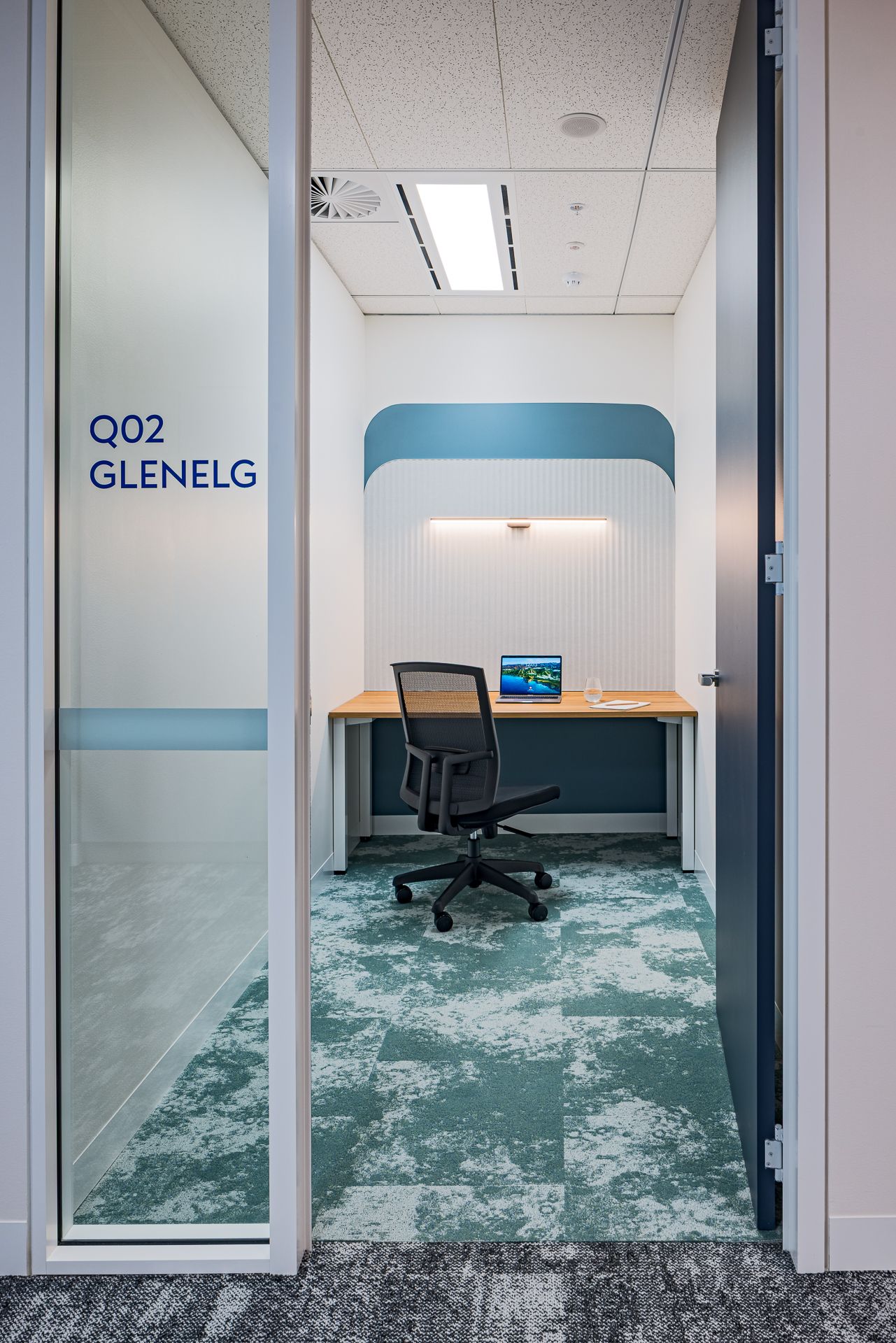 Private office focus booth labelled Q02 Glenelg with a desk, ergonomic black chair, monitor and green patterned carpet.