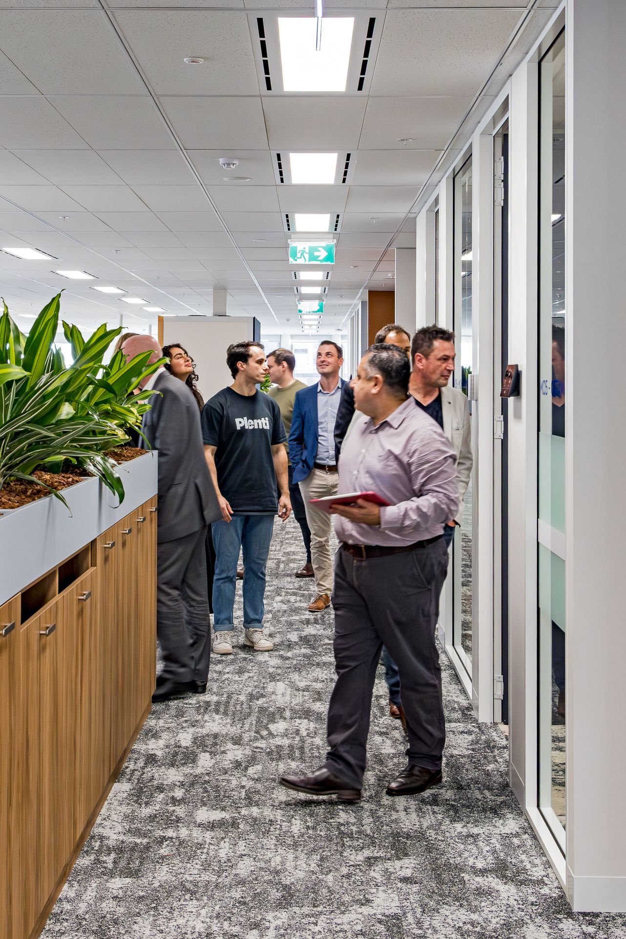 A group of colleagues gather in an office corridor beside timber planters and glass meeting rooms while one man holds a red folder.