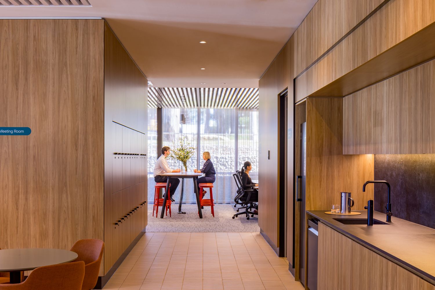 Colleagues chat at a high table in a bright office kitchenette with timber cabinetry, a stainless sink and red stools by large windows.