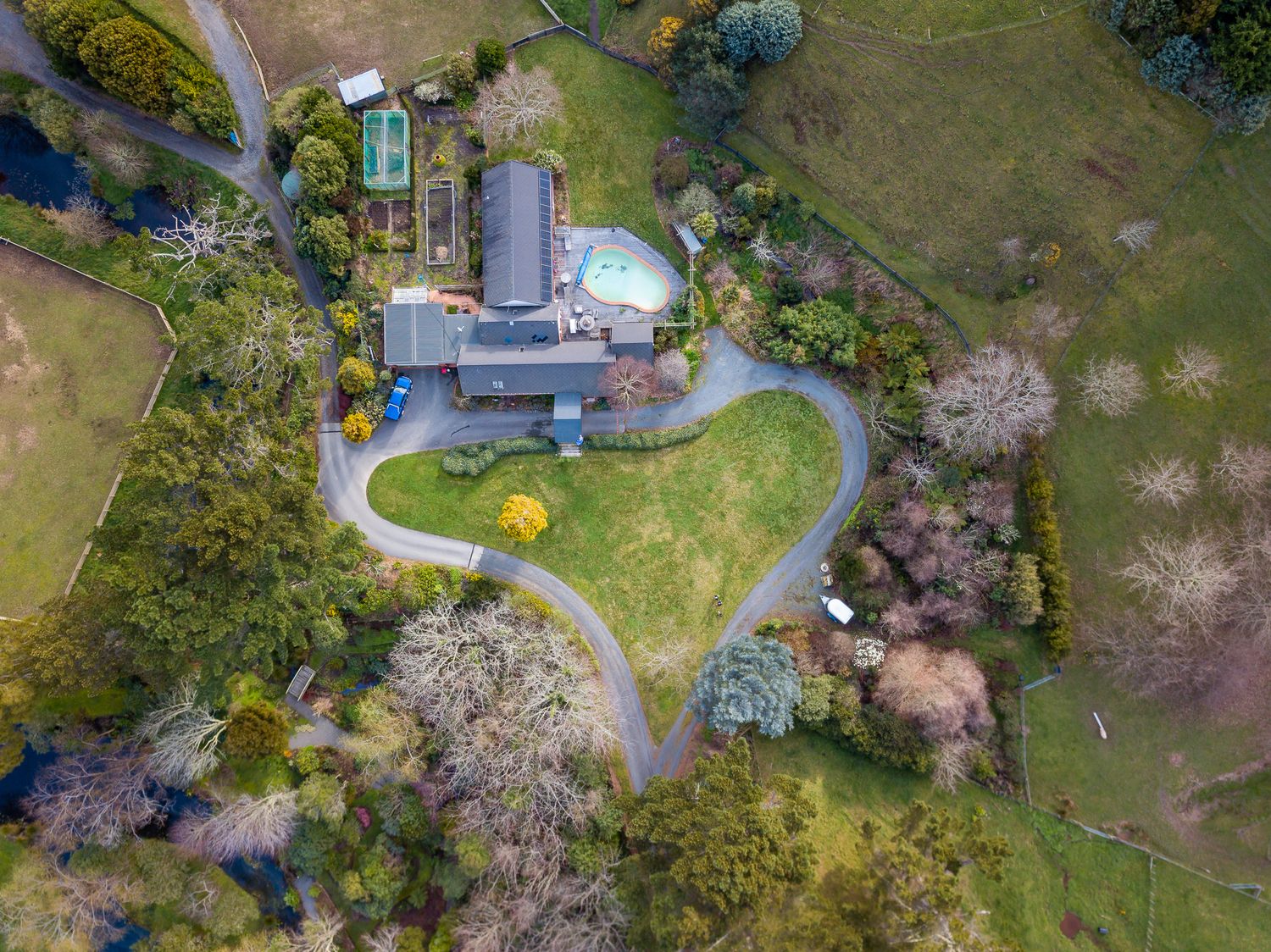 Aerial view of a countryside house with curved driveway, lawn, pool and surrounding trees and fields.