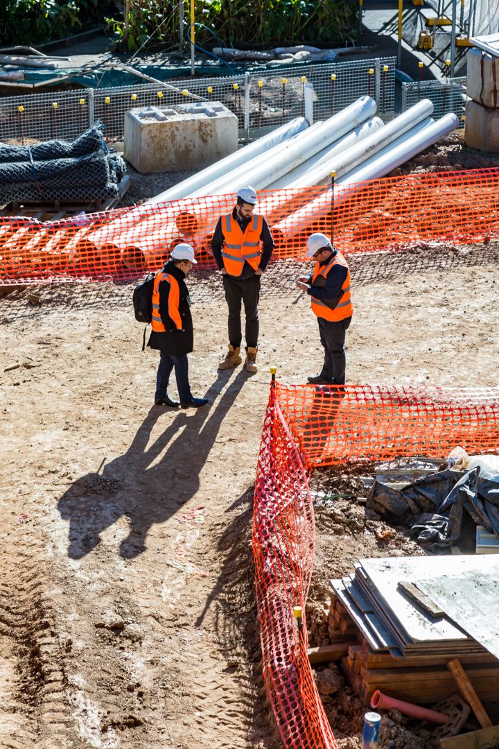Three workers in orange hi-vis vests and hard hats discuss plans at a construction site surrounded by orange safety fencing and large pipes.