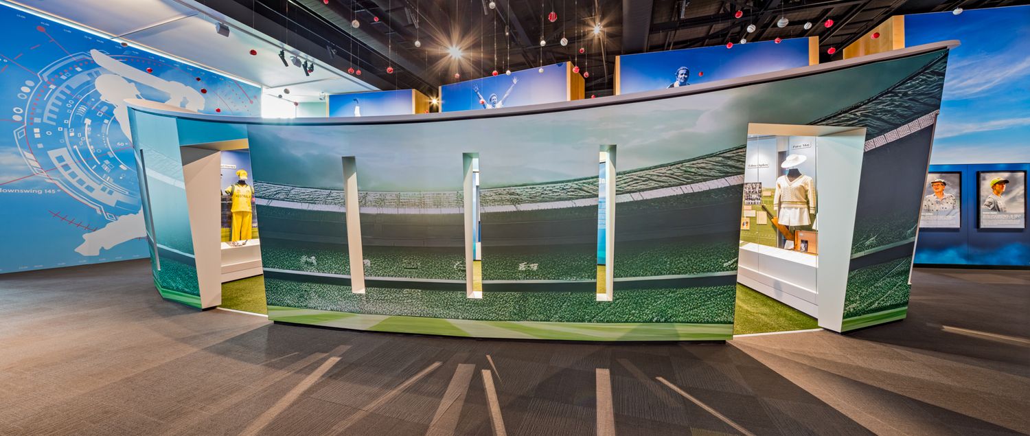 Curved museum exhibit wall printed with a cricket ground scene, with visitors viewing displays inside a modern gallery space.