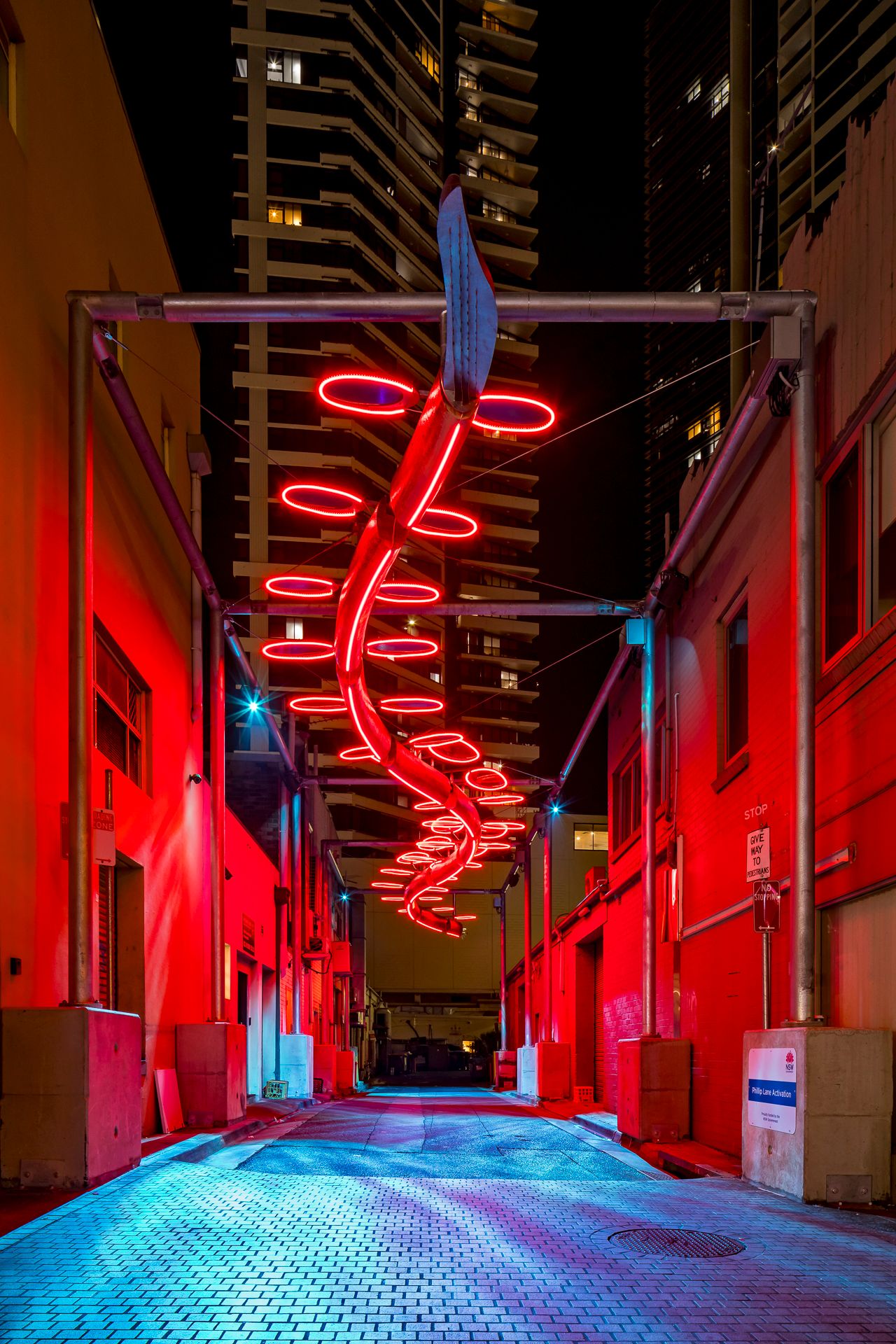 A red neon spiral light installation illuminates a narrow urban alley in vivid crimson tones after dark.