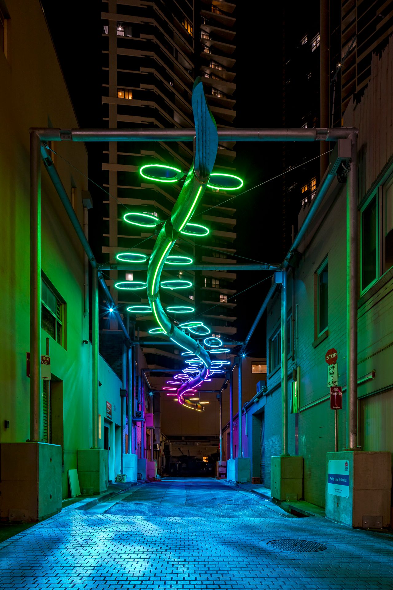 A green neon spiral art installation glows over a blue-lit cobblestone alley with a high-rise towering behind.