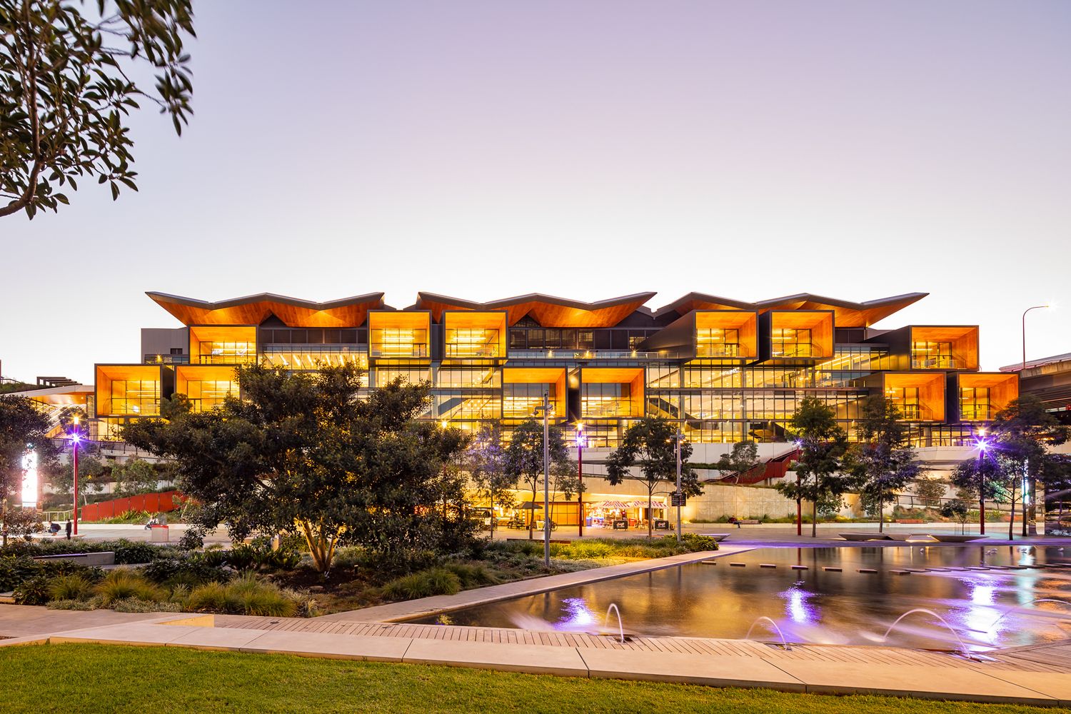 Illuminated convention center glowing orange and gold at twilight, with a reflective pool and trees in the foreground.