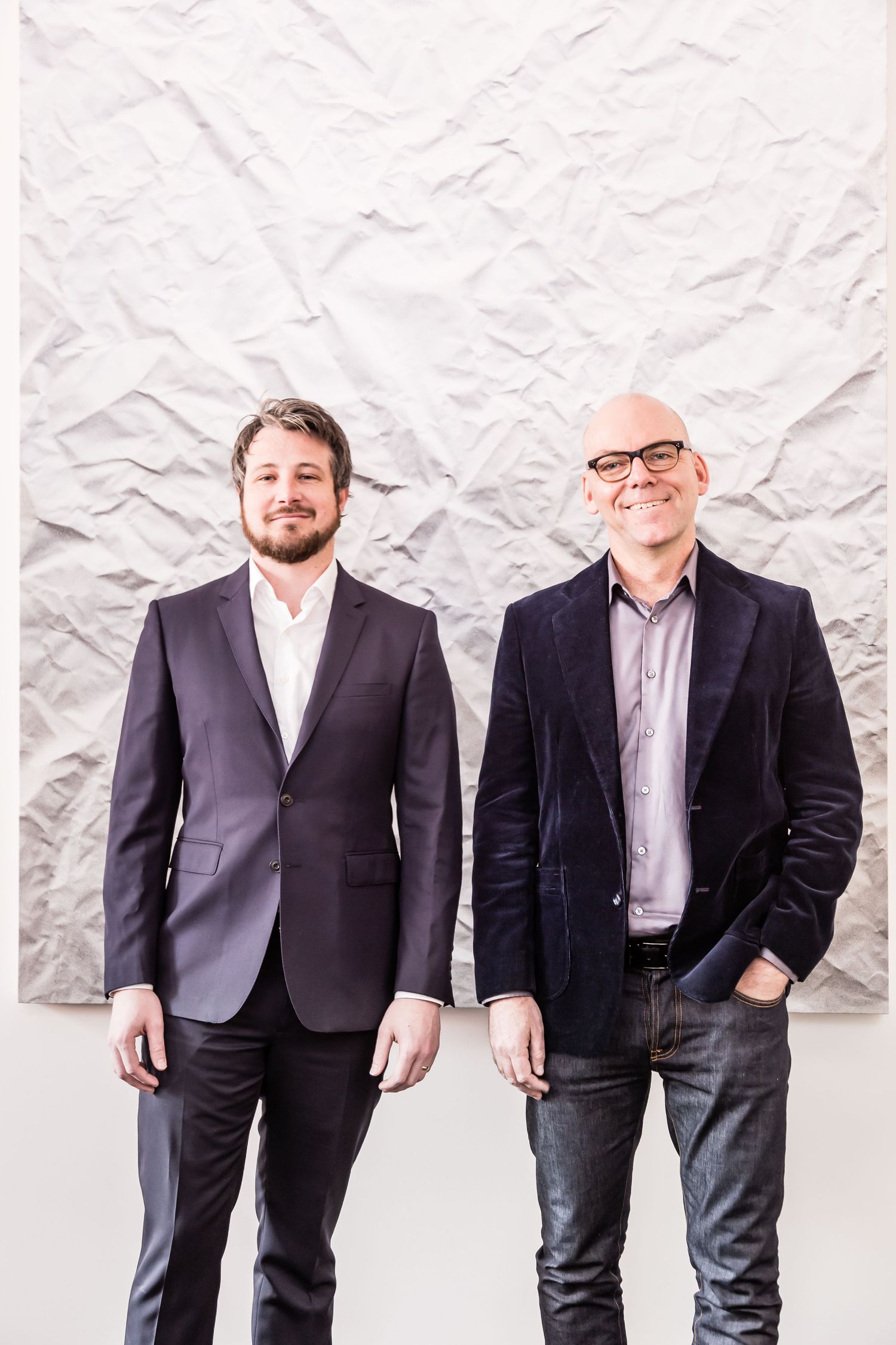 Two smiling men in blazers stand side by side against a textured white crumpled paper wall in a stylish portrait.