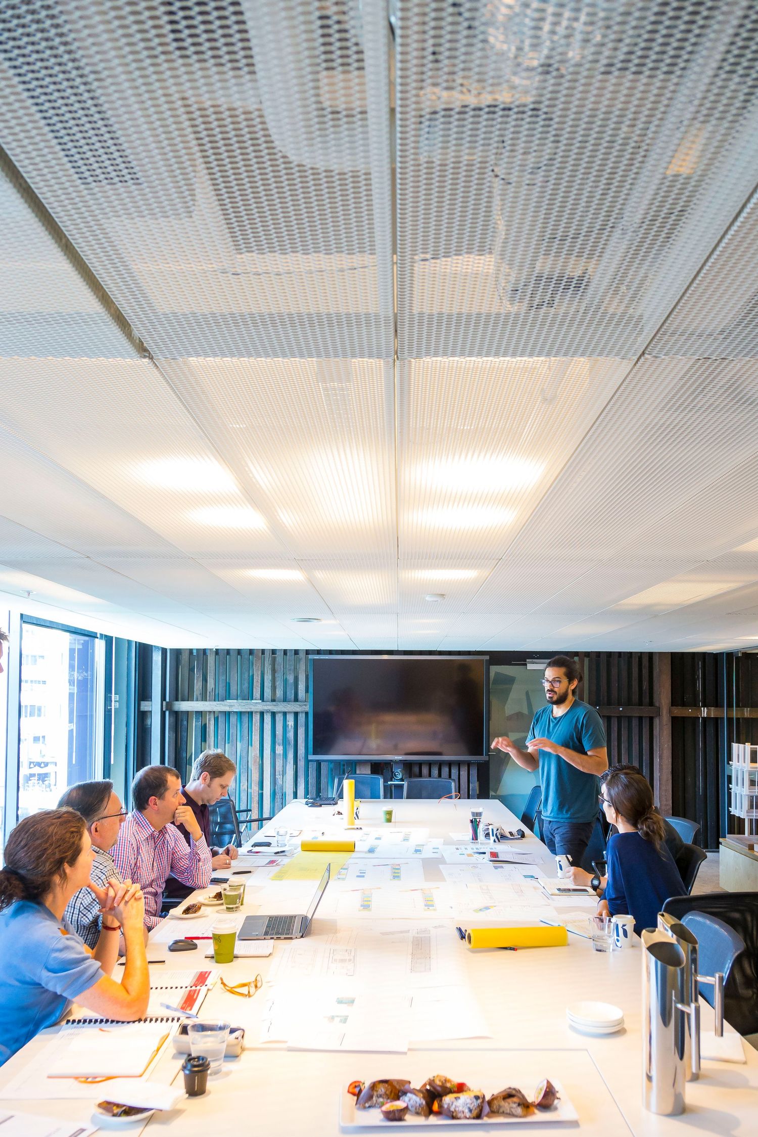 Team meeting in a modern conference room as a bbearded man presents to colleagues seated at a long table with documents and a wall screen.
