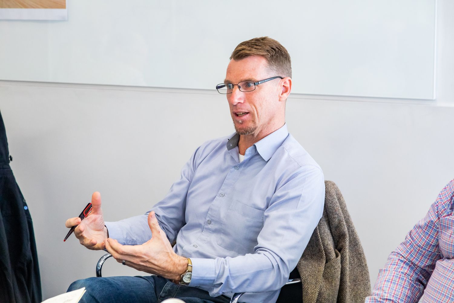 A man in glasses and a light blue shirt gestures with his hands while speaking during a discussion in a bright meeting room.