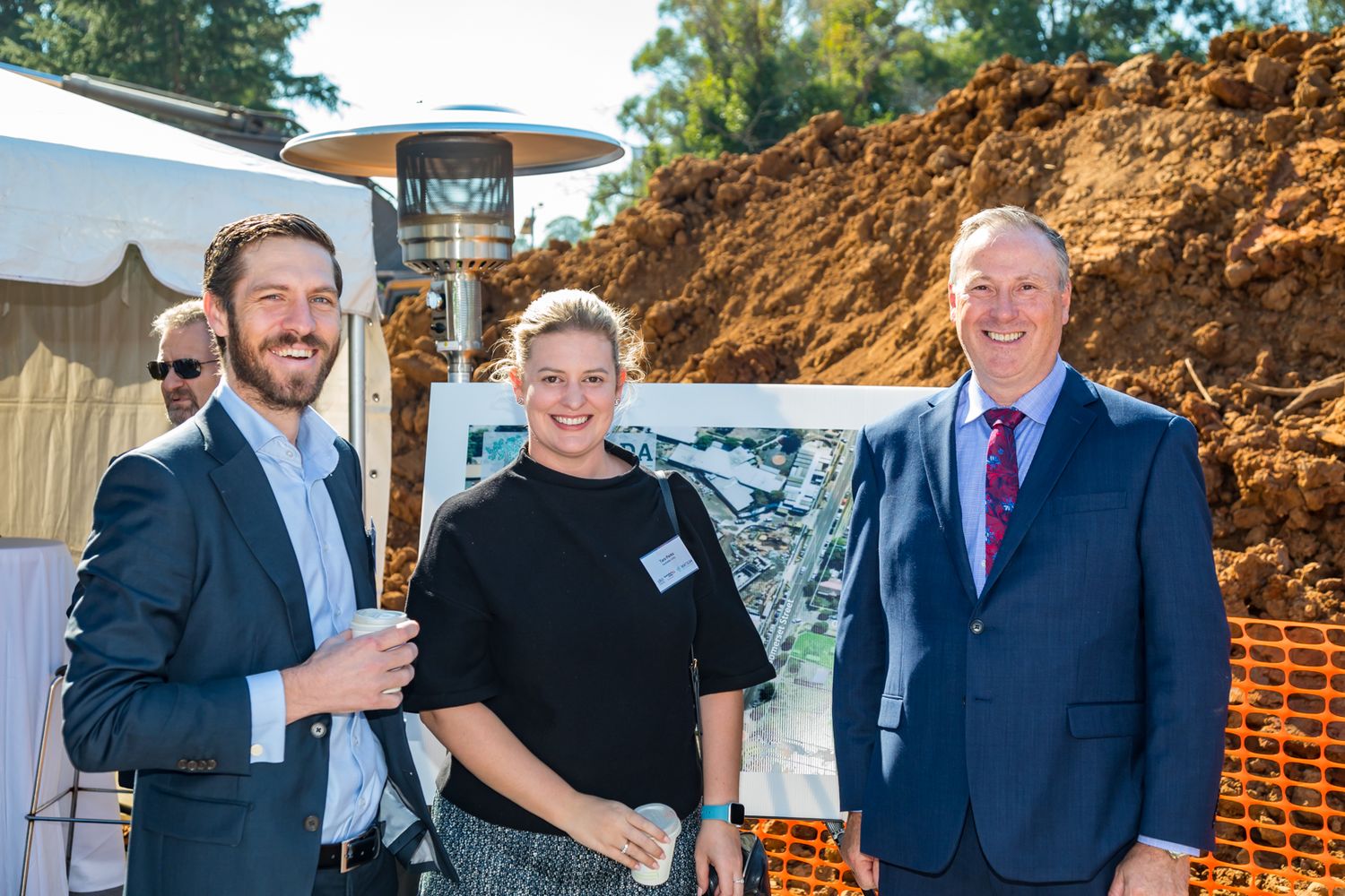 Two men in suits and a woman in black smile together at an outdoor groundbreaking event beside a mound of dirt and a tent.
