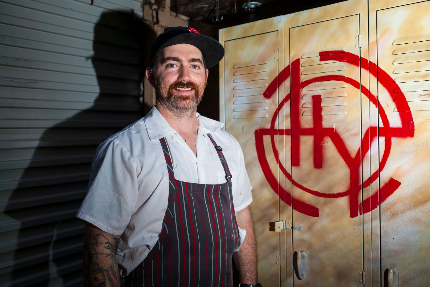 Bearded tattooed chef in a cap and striped apron smiles beside a metal locker marked with a red circular logo.