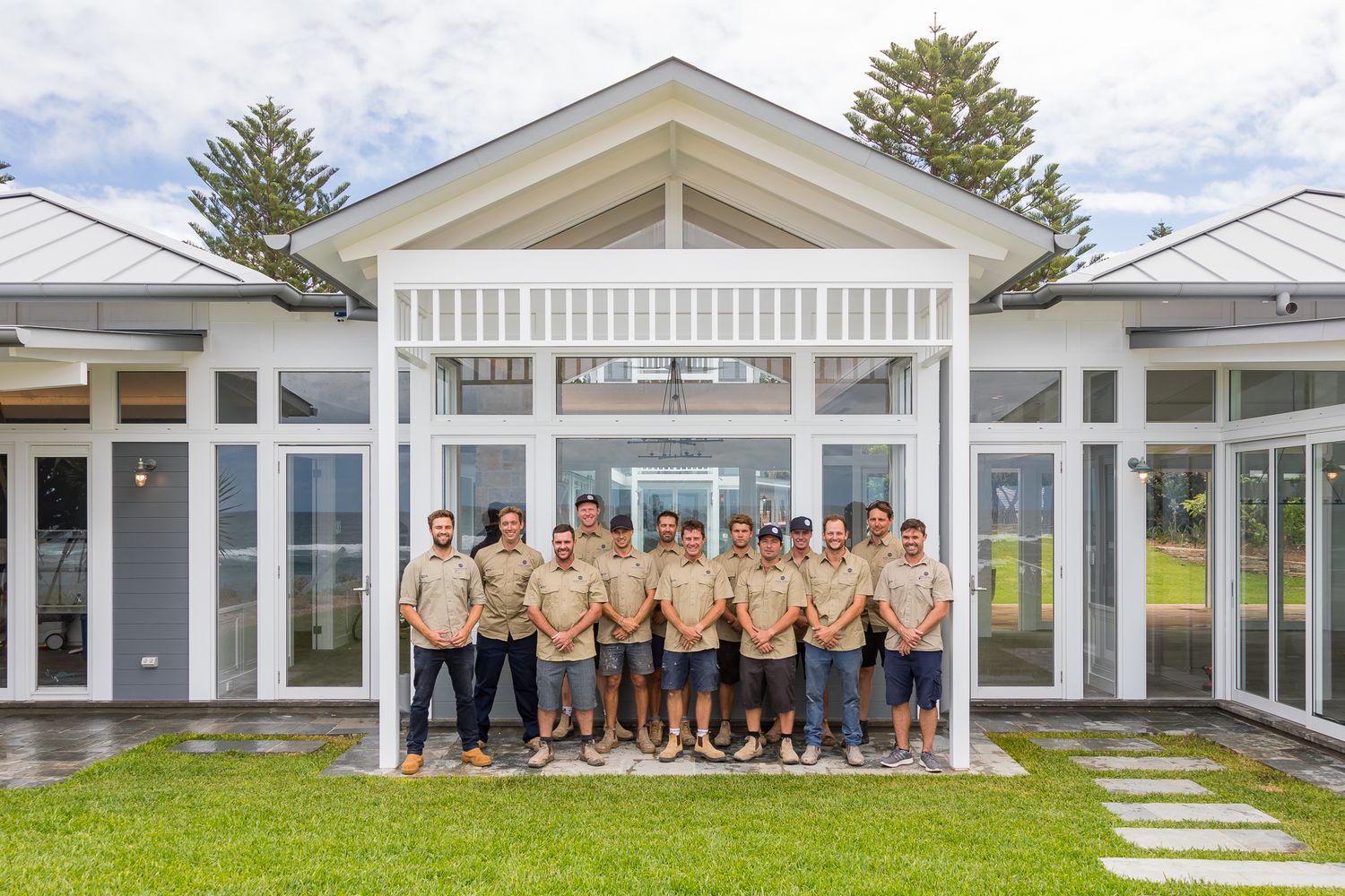 A group of men in matching tan shirts stand together in front of a modern white single-storey building with a lawn.