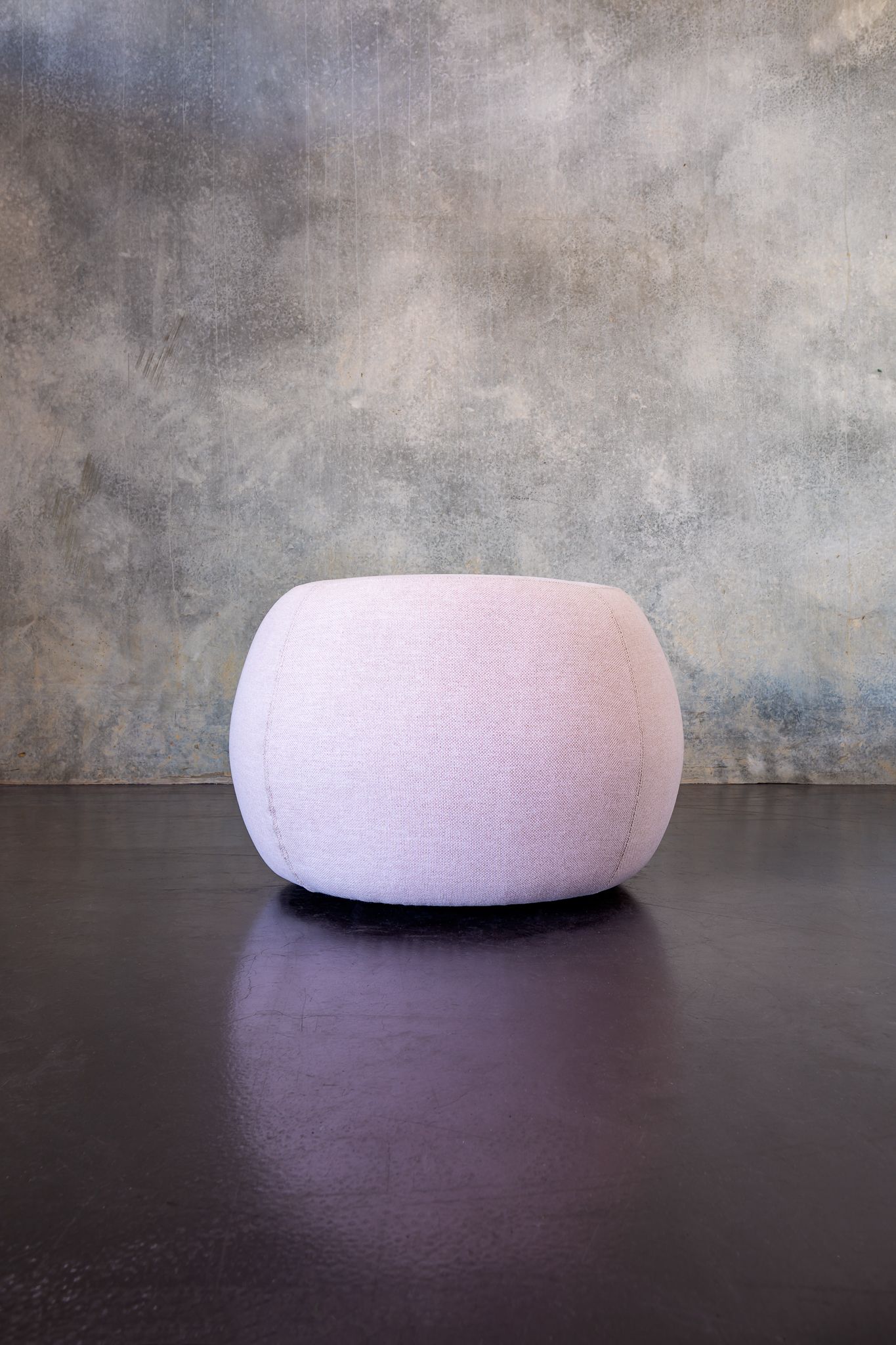 Round blush pink upholstered pouf ottoman on a dark glossy floor against a textured concrete-look wall.