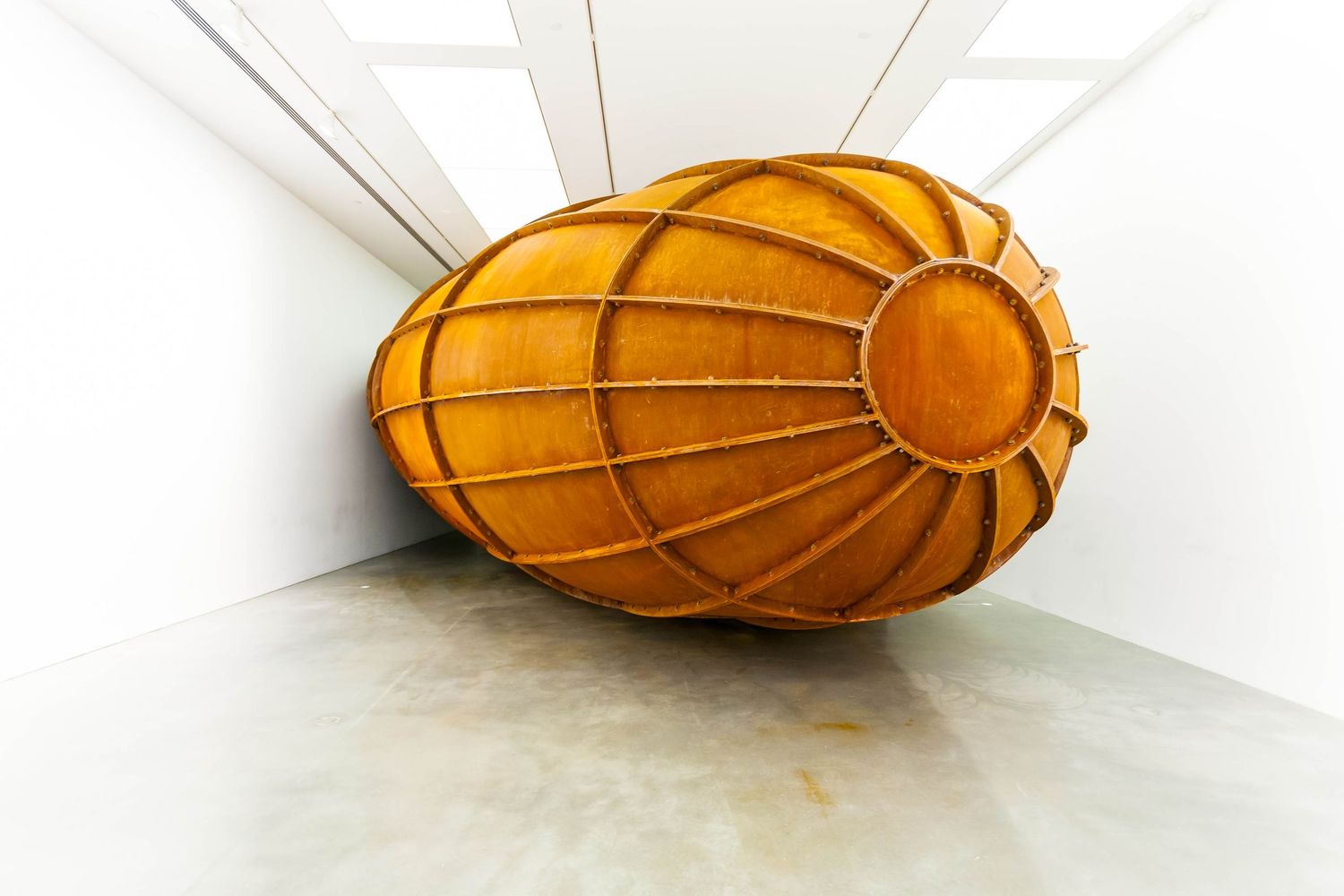 A large rusted steel sculpture shaped like an oval capsule with ribbed supports sits inside a bright white gallery space.