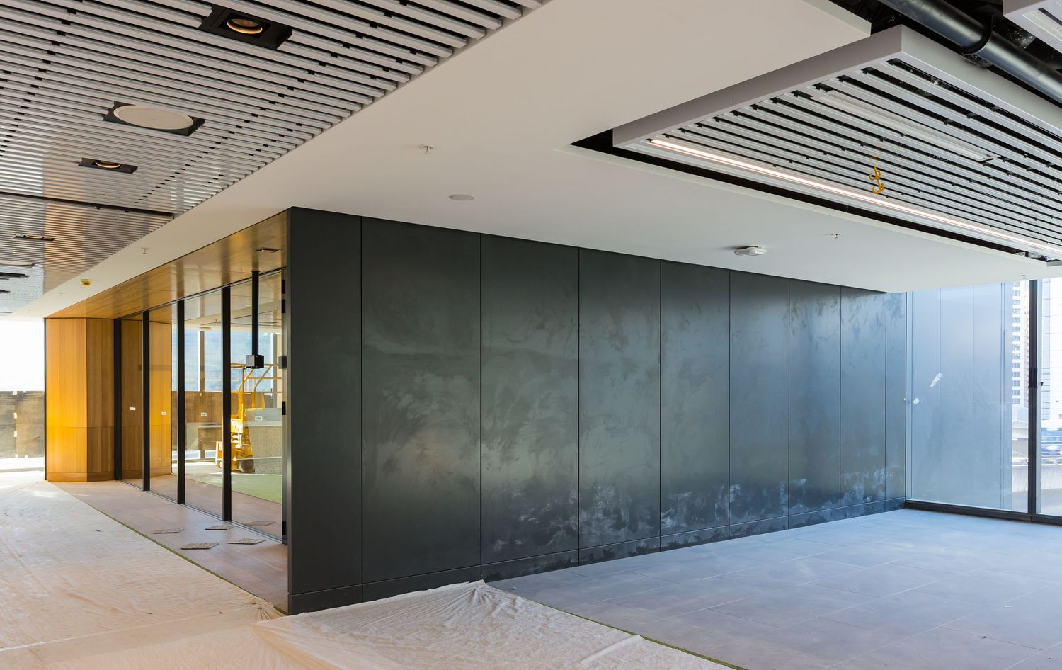 Modern office corridor with dark panelled wall, glass-fronted meeting room, slatted timber ceiling, and bright floor-to-ceiling windows.