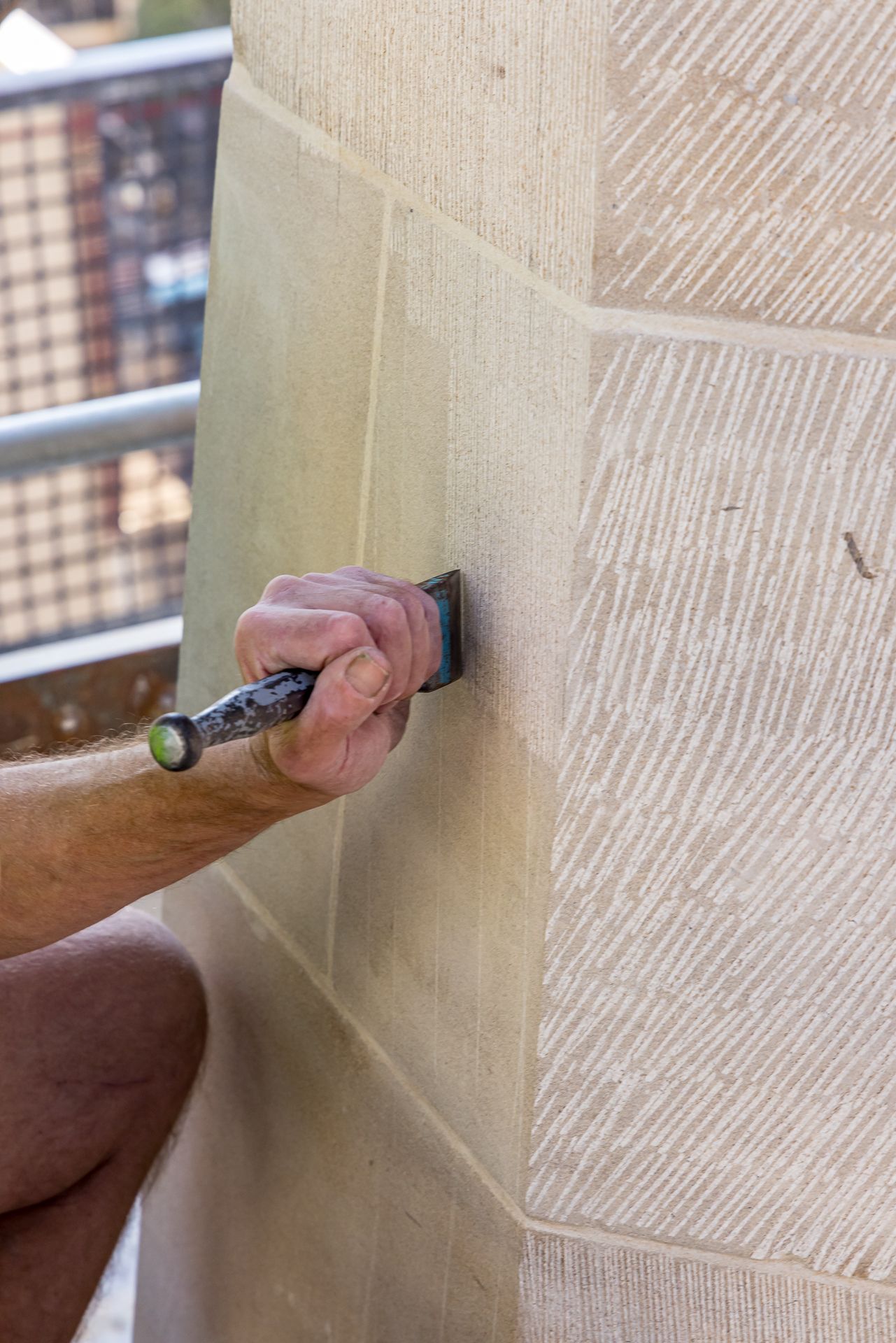 Hand holding a chisel carves a smooth edge into a pale sandstone block with textured tooled surface.