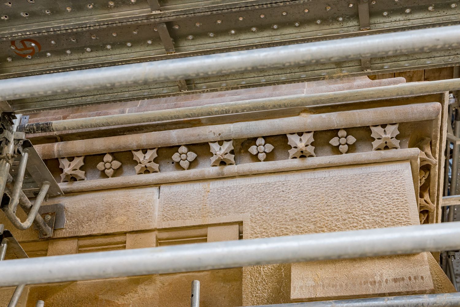 Row of carved stone rosettes along a sandstone cornice seen through metal scaffolding poles during building restoration.