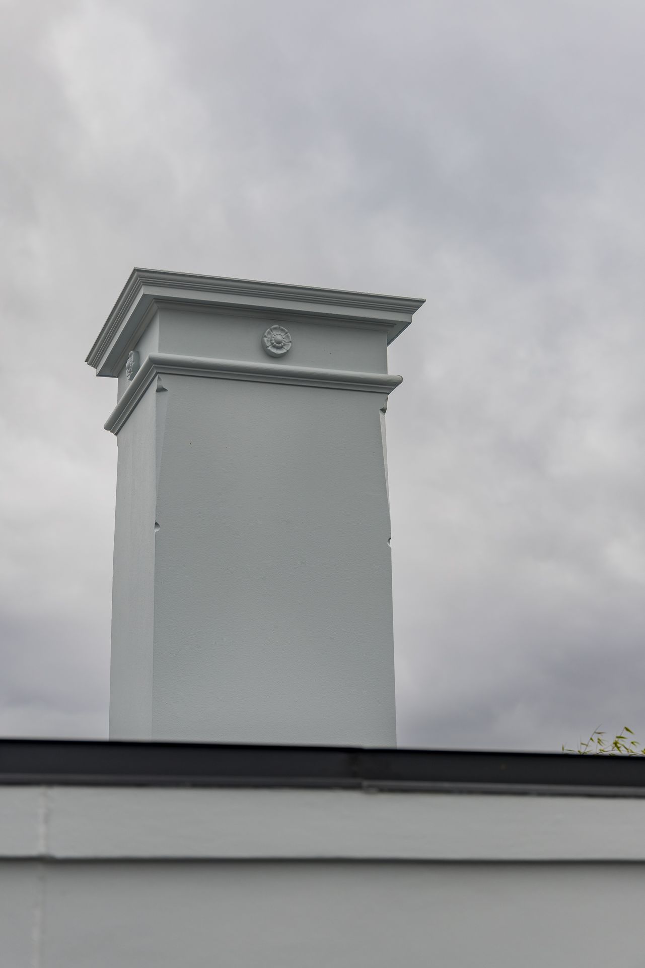 Freshly painted pale grey square chimney with decorative cornice and rosette detail against an overcast sky.