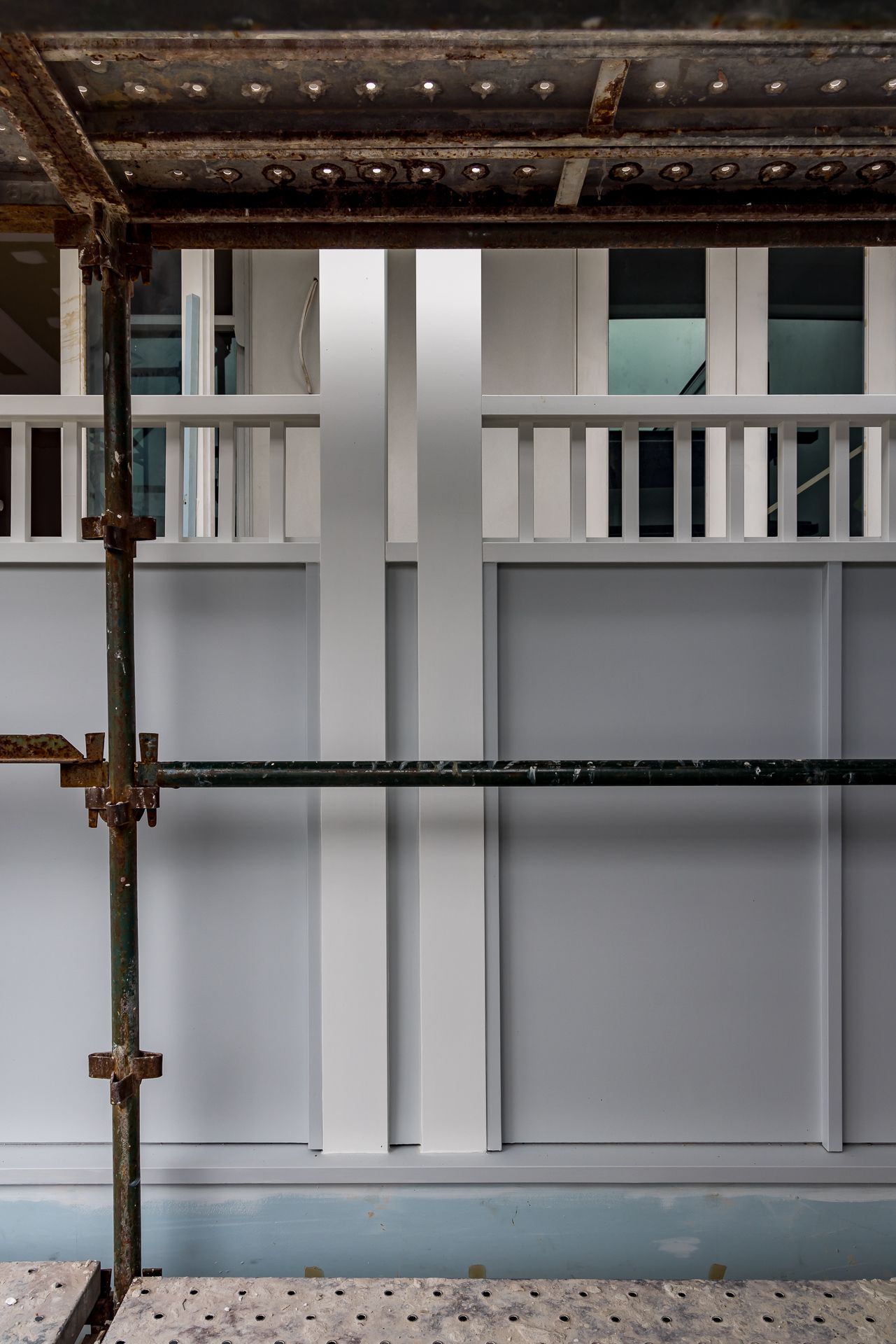 White painted panelled wall with balustrade detail seen behind rusted construction scaffolding inside a renovation site.