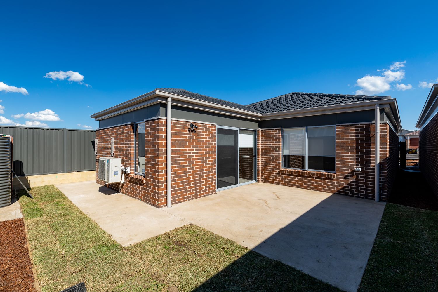 Newly built single-storey brick home with a dark tiled roof, sliding glass door and a concrete patio beside a freshly laid lawn.