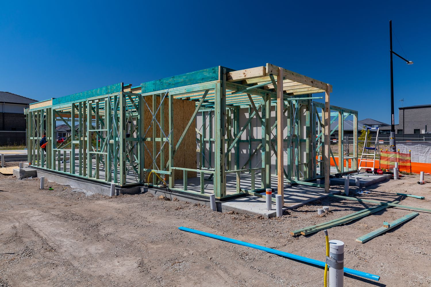 Green timber wall and roof framing of a new home under construction on a cleared building site beneath a clear blue sky.