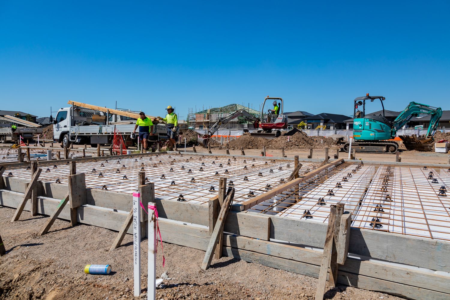 Construction workers in high-visibility clothing beside a concrete slab foundation with timber formwork and excavators nearby.