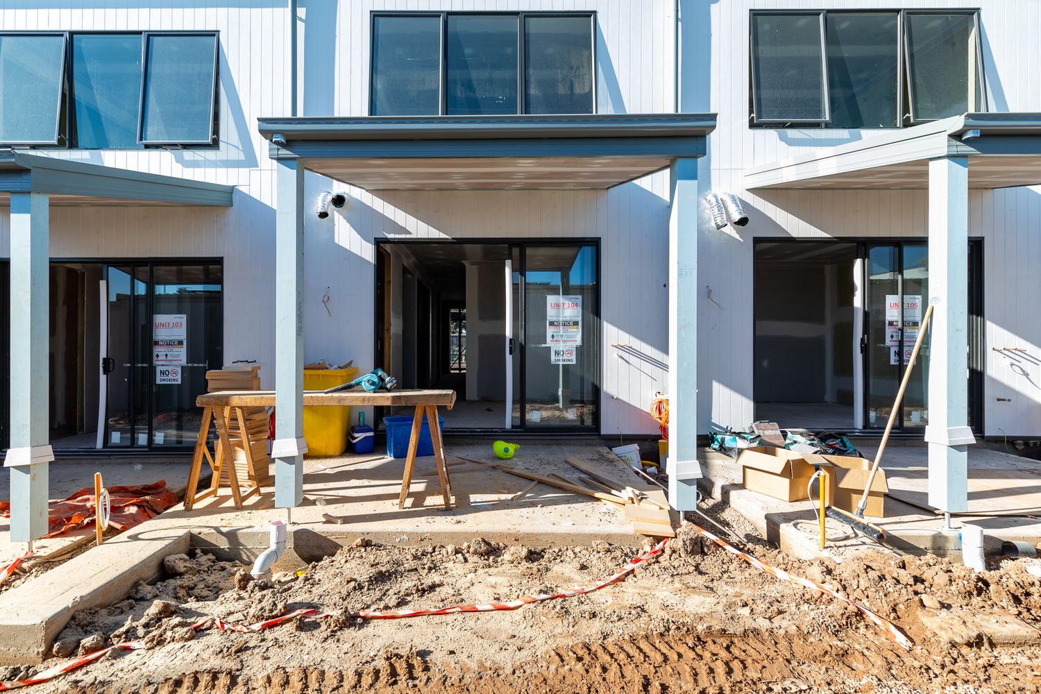 Front of a new multi-storey townhouse development under construction, with timber trestles, large windows and a muddy building site.