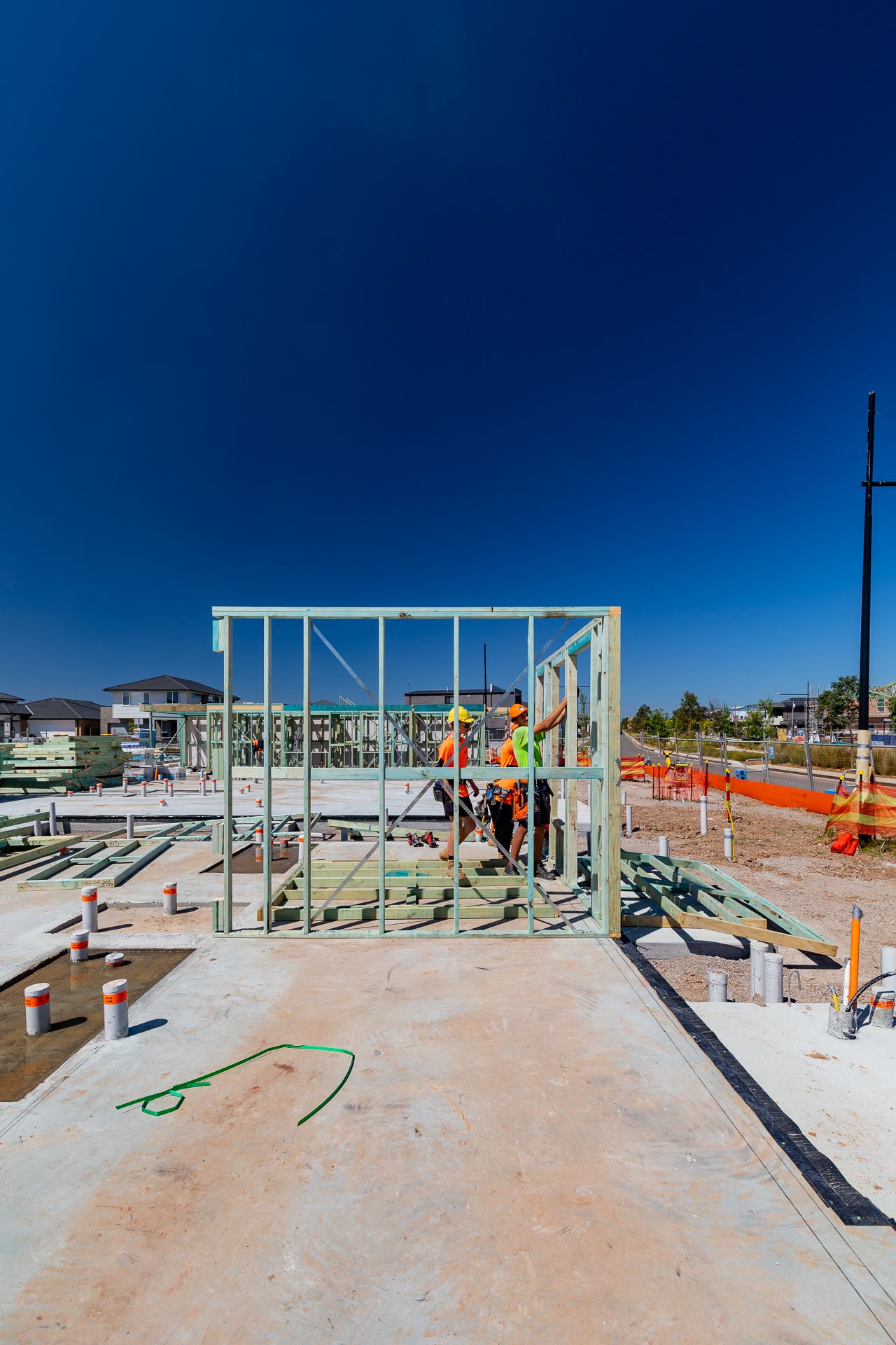Construction workers in hi-vis gear assemble a timber wall frame on a concrete slab at a new housing development site.