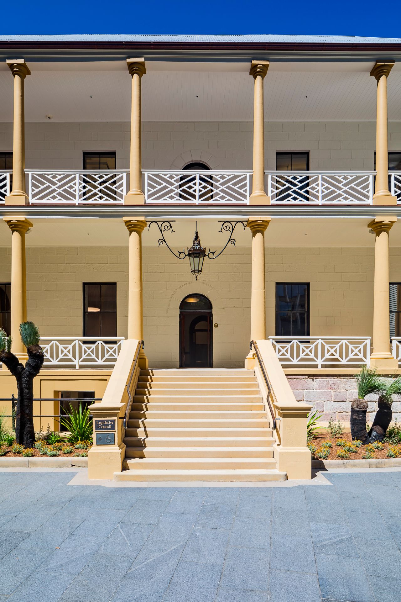 Symmetrical two-storey colonial building with cream columns, lattice balustrades, a central arched doorway, and a wide stone staircase.