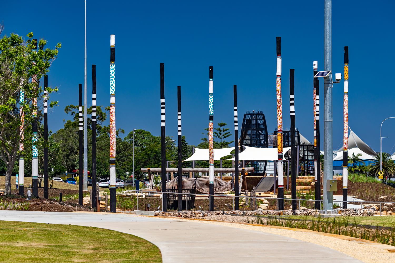 Rows of tall black poles with colourful painted bands line a curved path leading to a park playground with shade sails.
