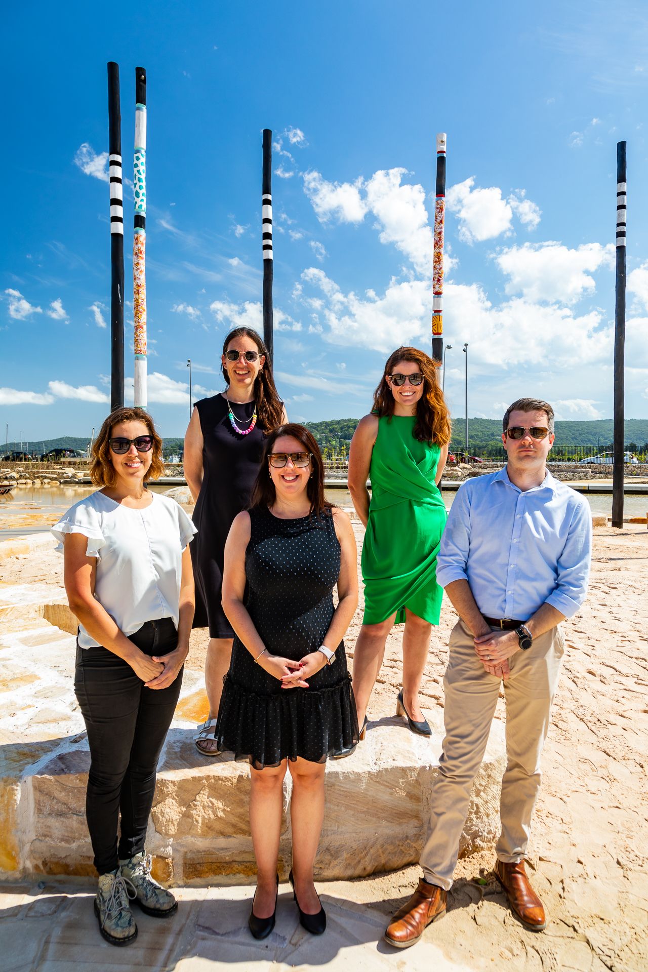 Five smiling people pose together on sandstone steps beside tall painted poles at a sunny waterfront park under a blue sky.