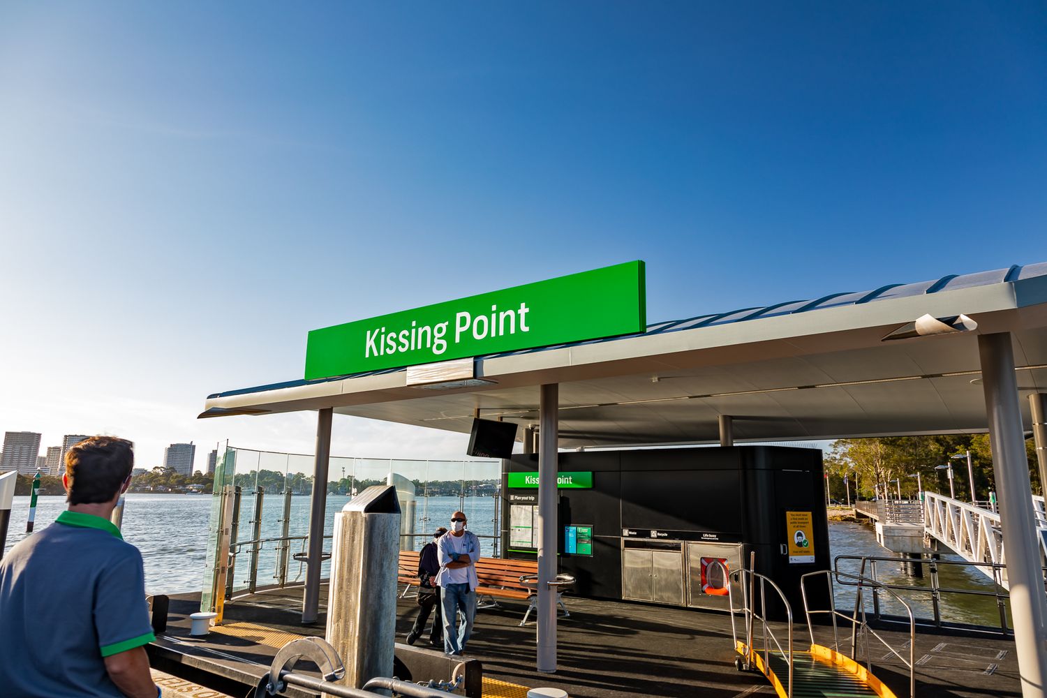 Two passengers stand on the Kissing Point ferry wharf platform below the green station sign on a bright sunny day.