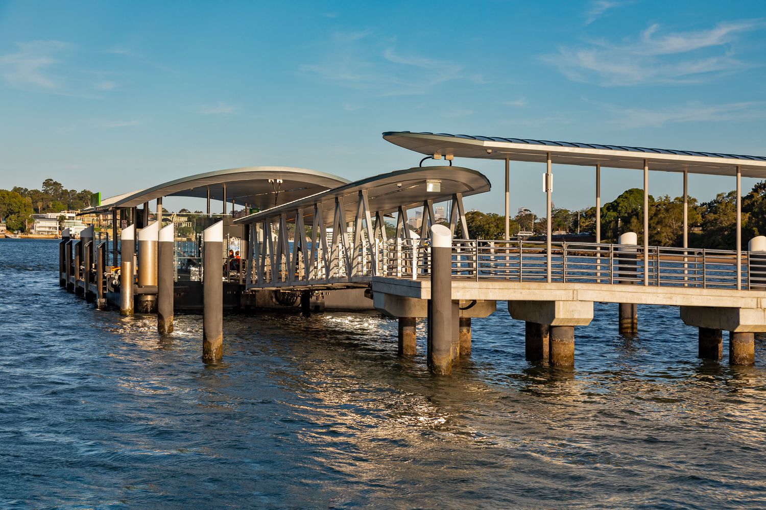 Curved canopy roof and steel posts of a modern ferry wharf extending over calm blue water under a clear sky.