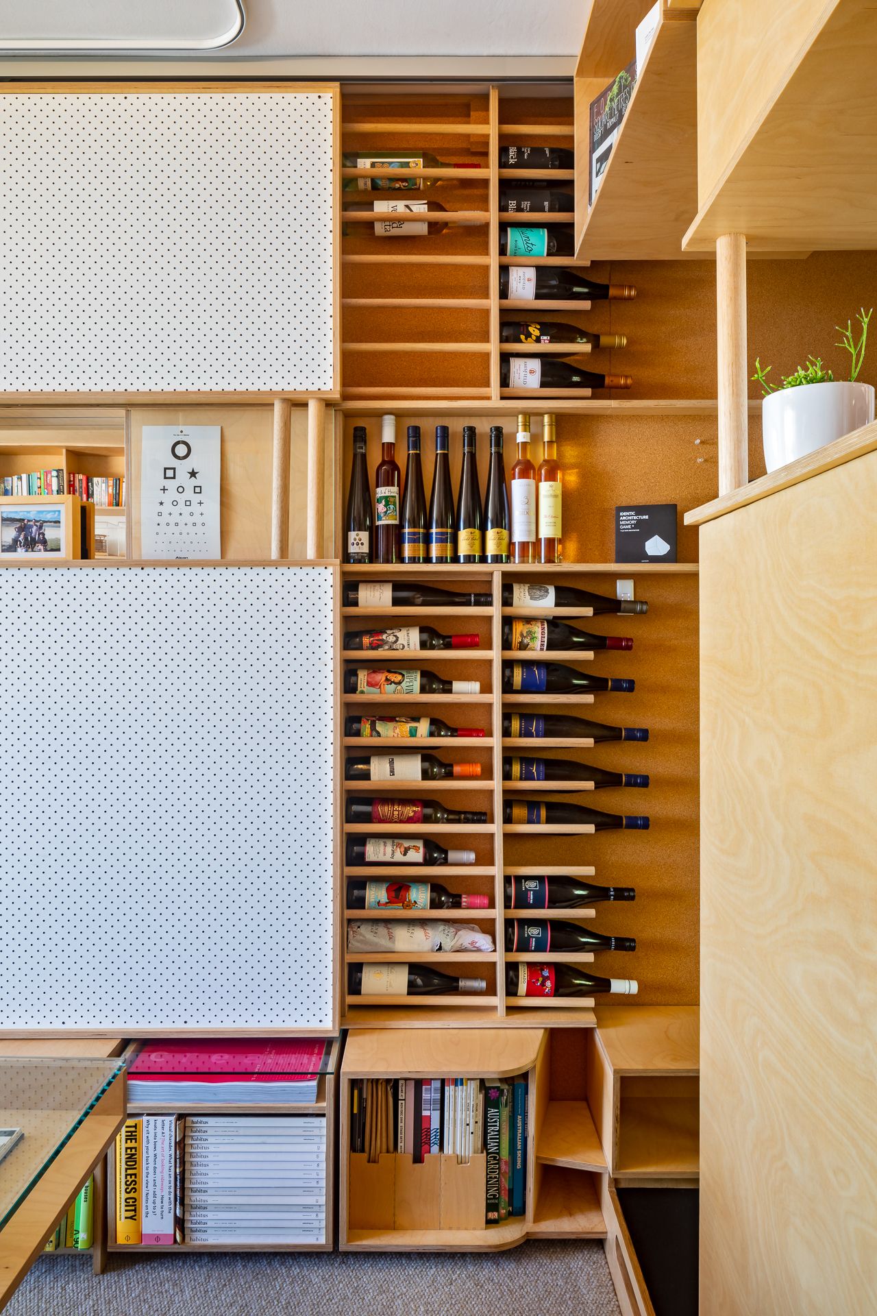 Plywood wall unit with angled wine bottle racks, pegboard panels, books and an eye chart in a compact apartment.