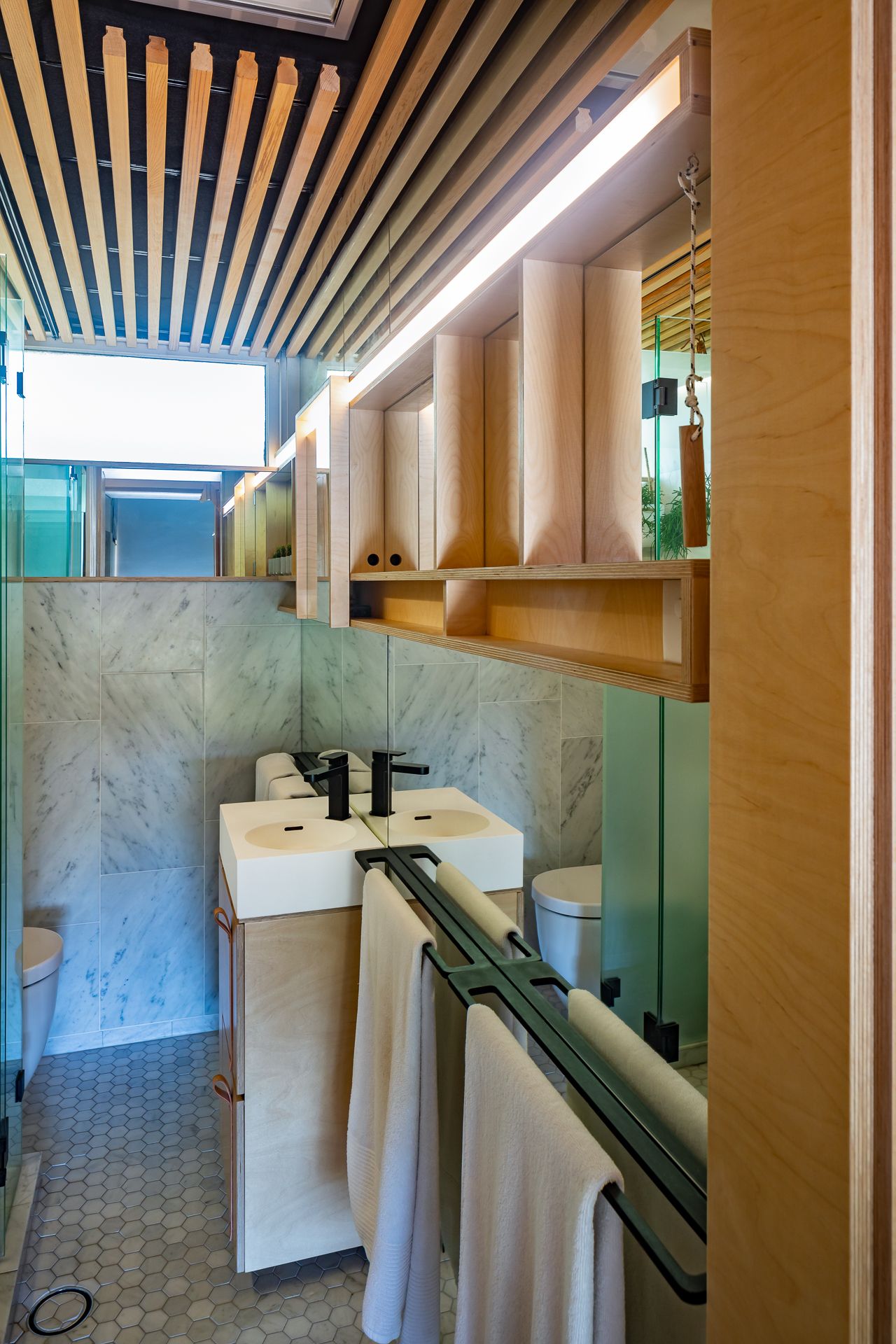 Narrow bathroom with marble walls, a white vanity sink, black fixtures, hanging towels, and a slatted timber ceiling.