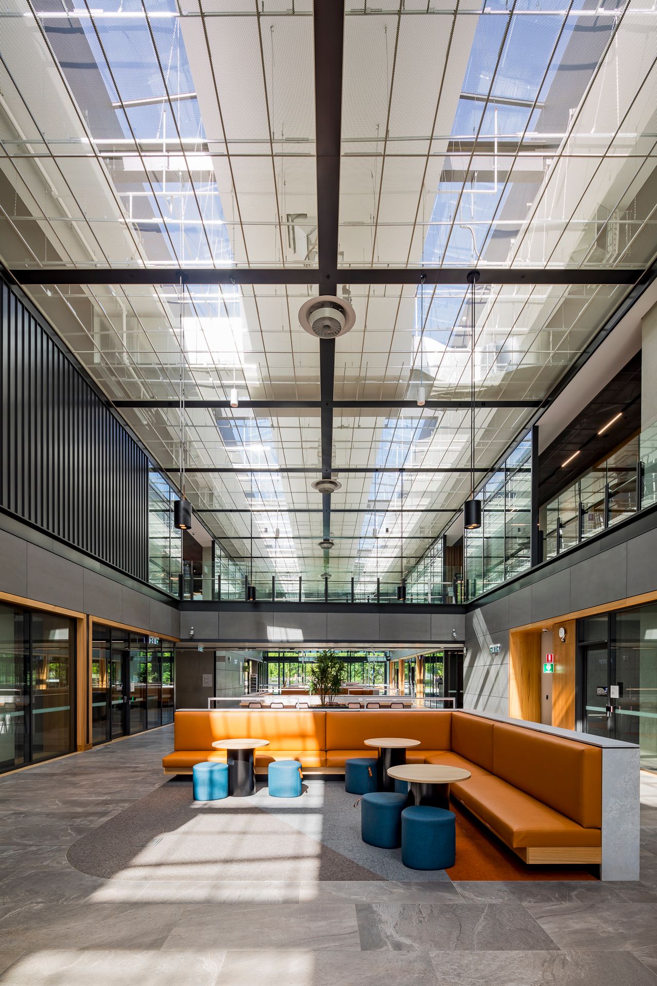 Sunny multi-level atrium with a glass skylight roof, orange banquette seating, round tables and teal stools.