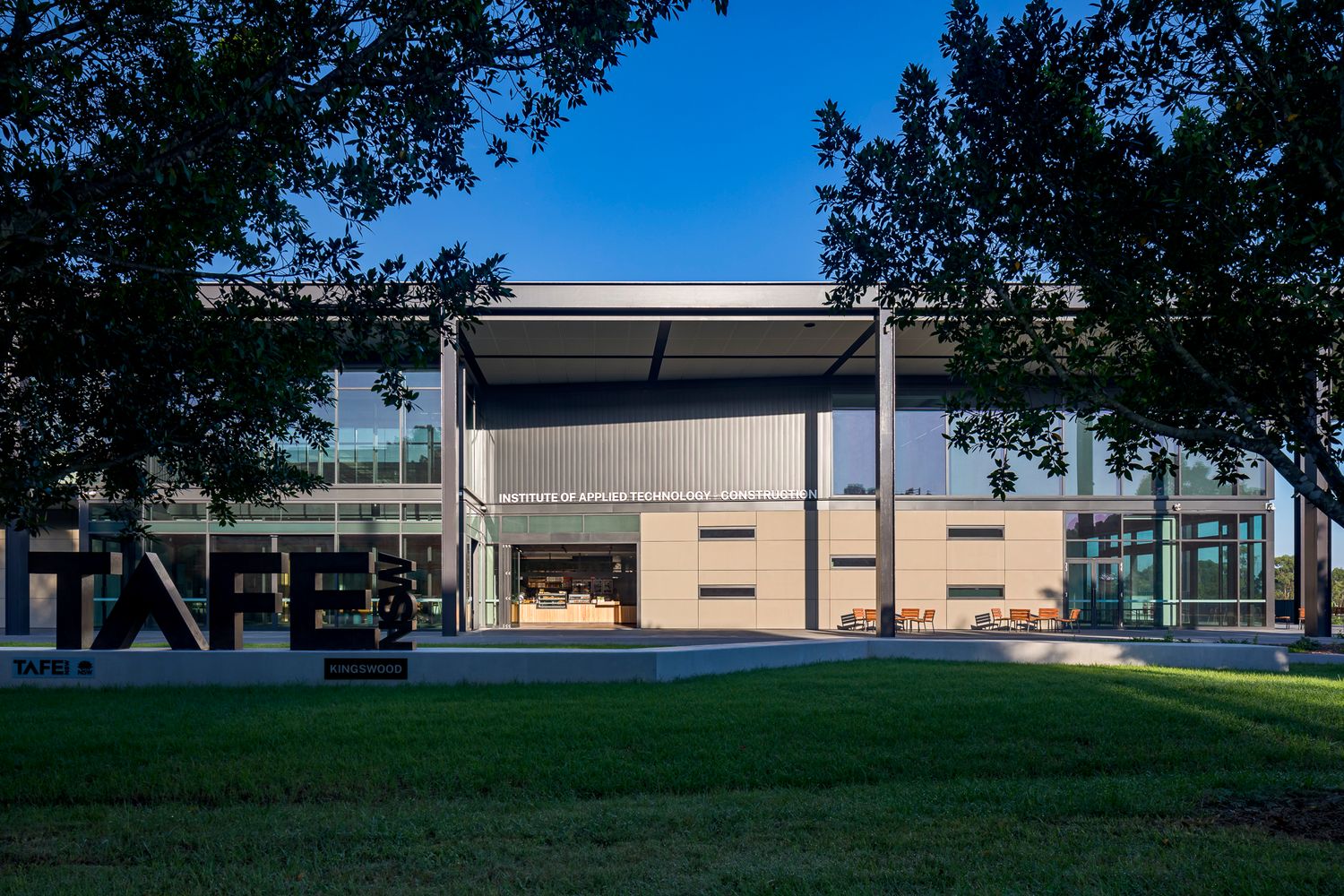 Entrance to a TAFE campus building with signage, steel frame and tall glass windows framed by tree branches and green lawn.