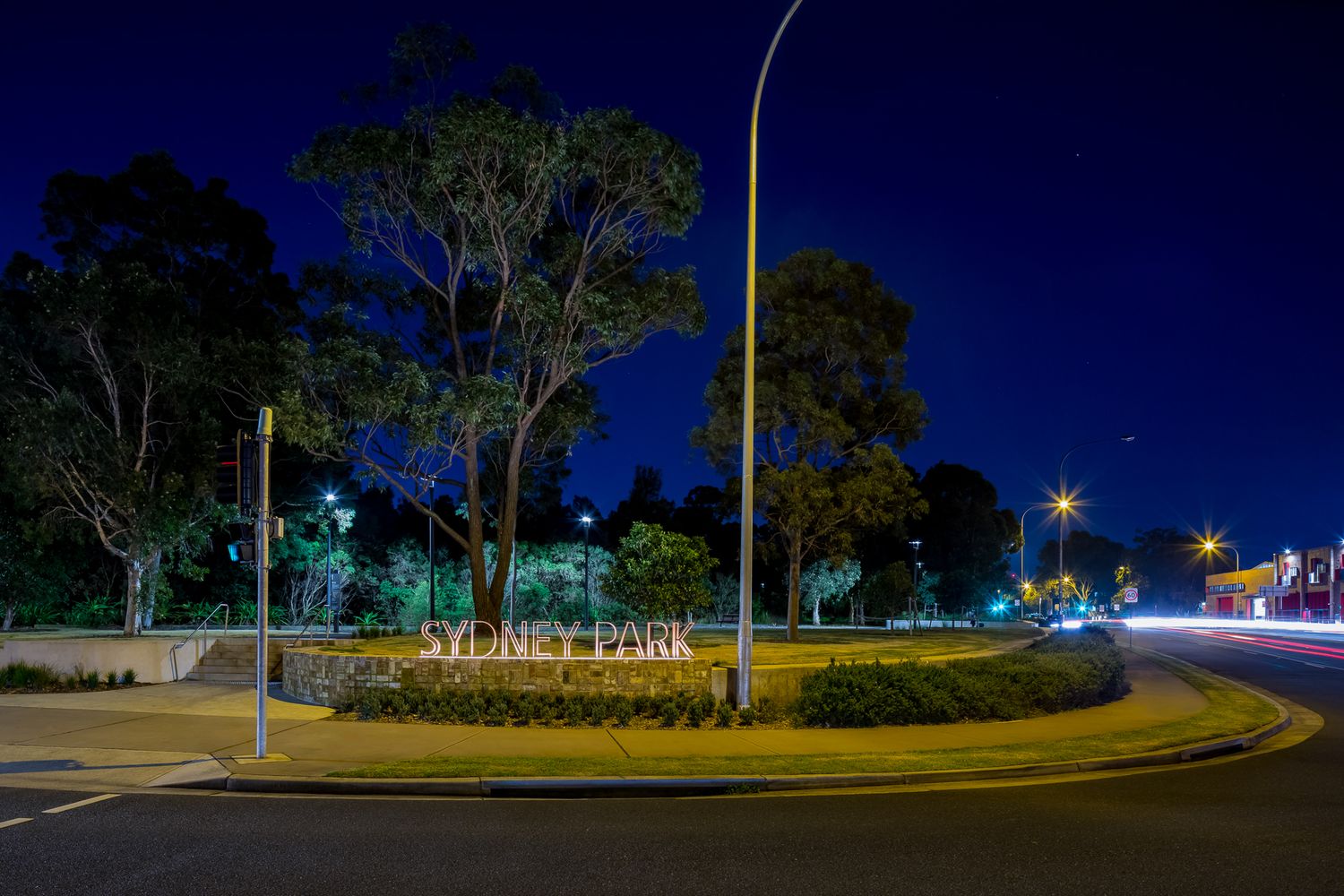 Sydney Park entrance sign lit at night beside a curving road, streetlights and silhouetted trees under a deep blue sky.