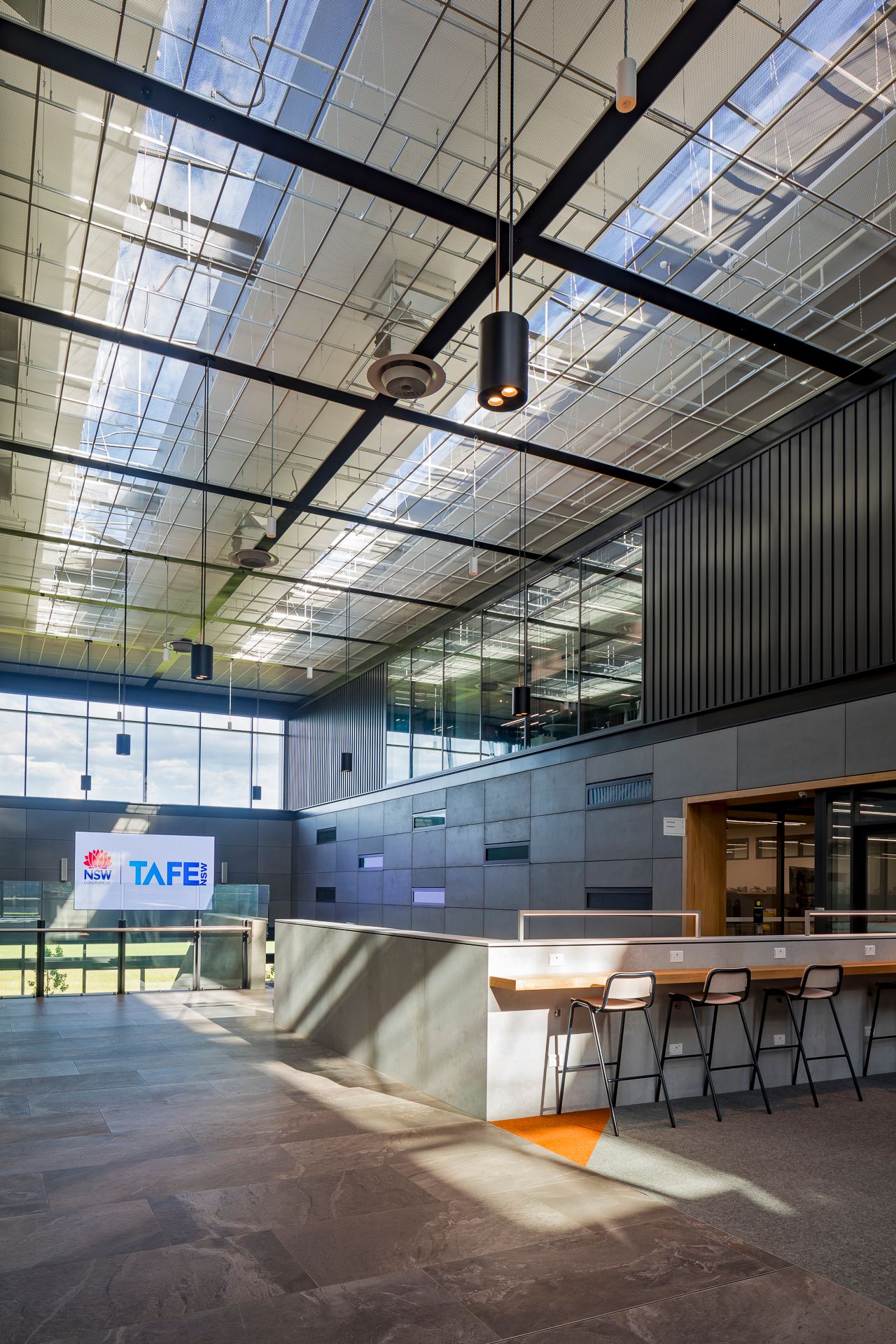 TAFE campus foyer with high skylit ceiling, branded signage, a reception counter with bar stools and dark cladding walls.