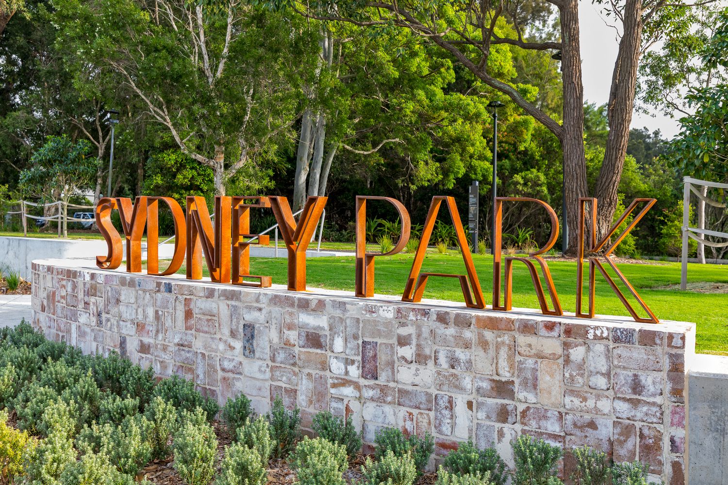 Visitors rest on a lawn beside a curved concrete seat wall and steps with metal handrails, with eucalyptus trees and brick buildings behind.