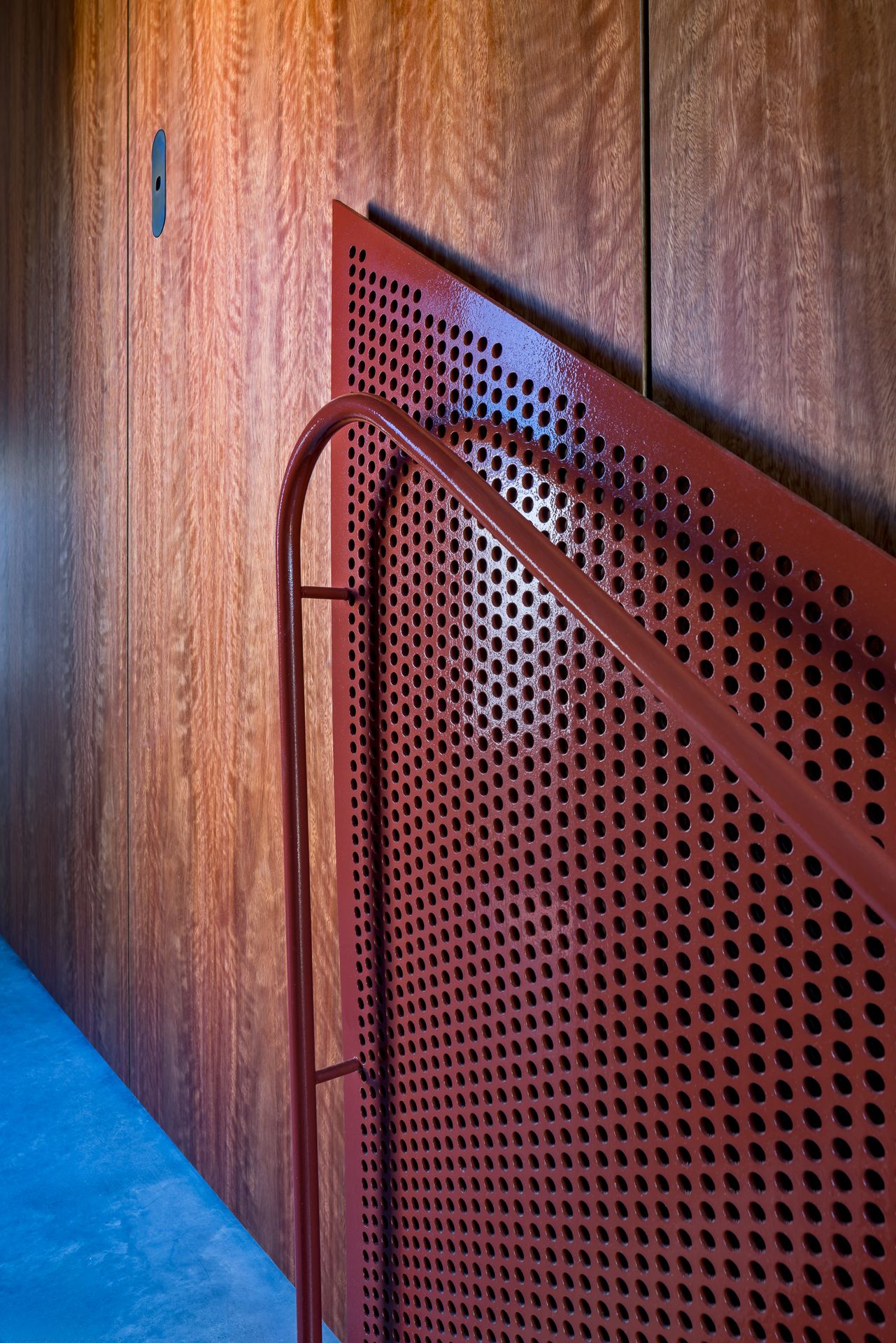Close-up of a red perforated metal stair balustrade with a tubular handrail against warm timber wall paneling and a concrete floor.