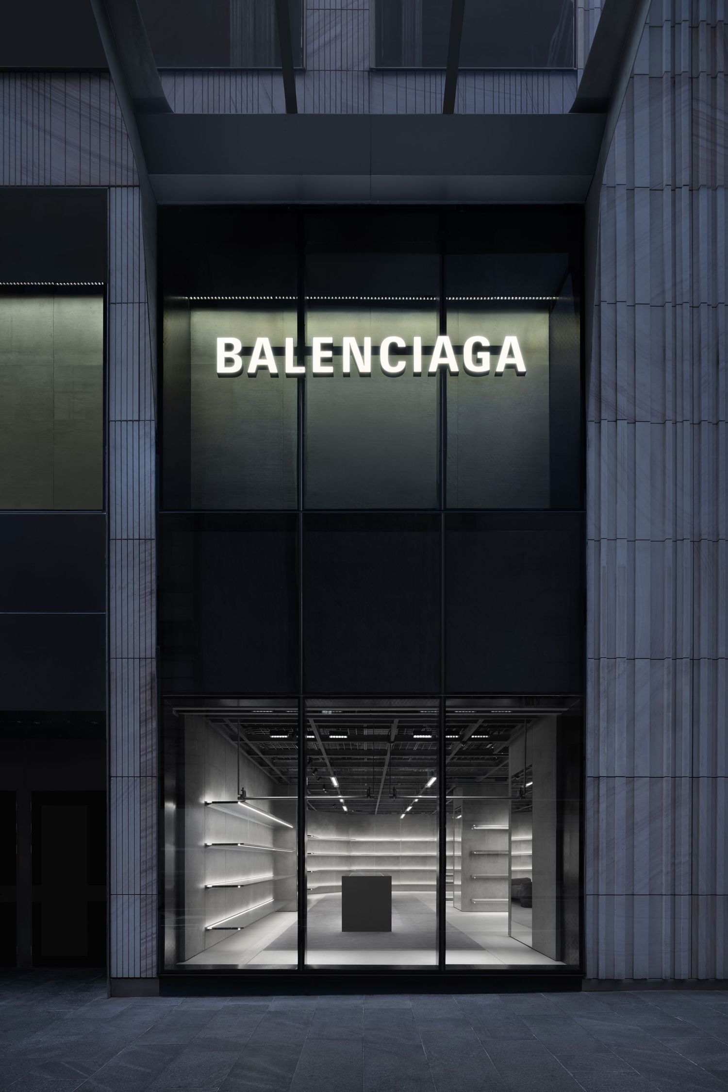 Balenciaga luxury boutique entrance with a backlit logo on a dark facade and a glowing, shelf-lined gray display interior.