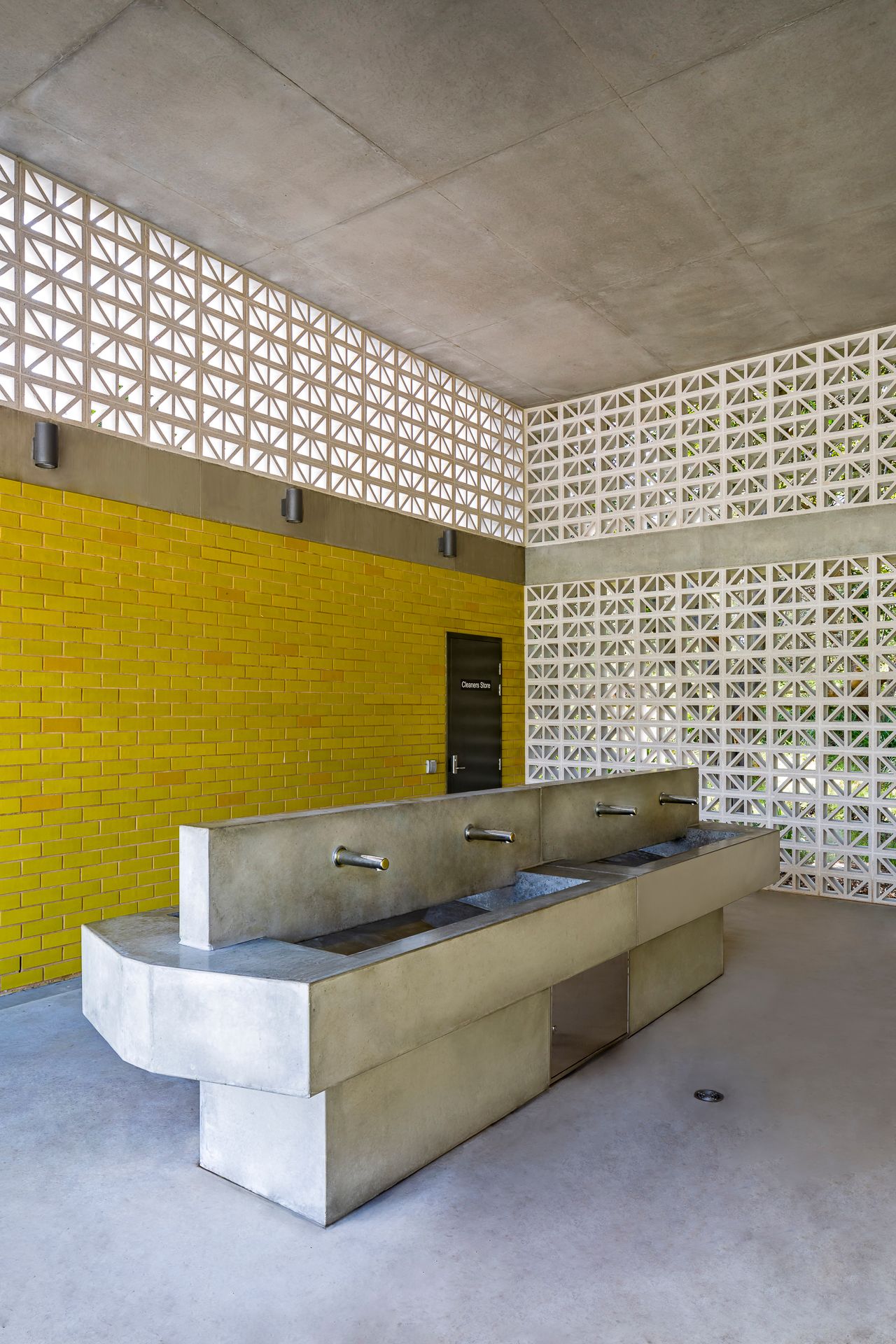 Public restroom interior with a bright yellow brick wall, concrete trough sink with metal taps and white decorative breeze block screens.