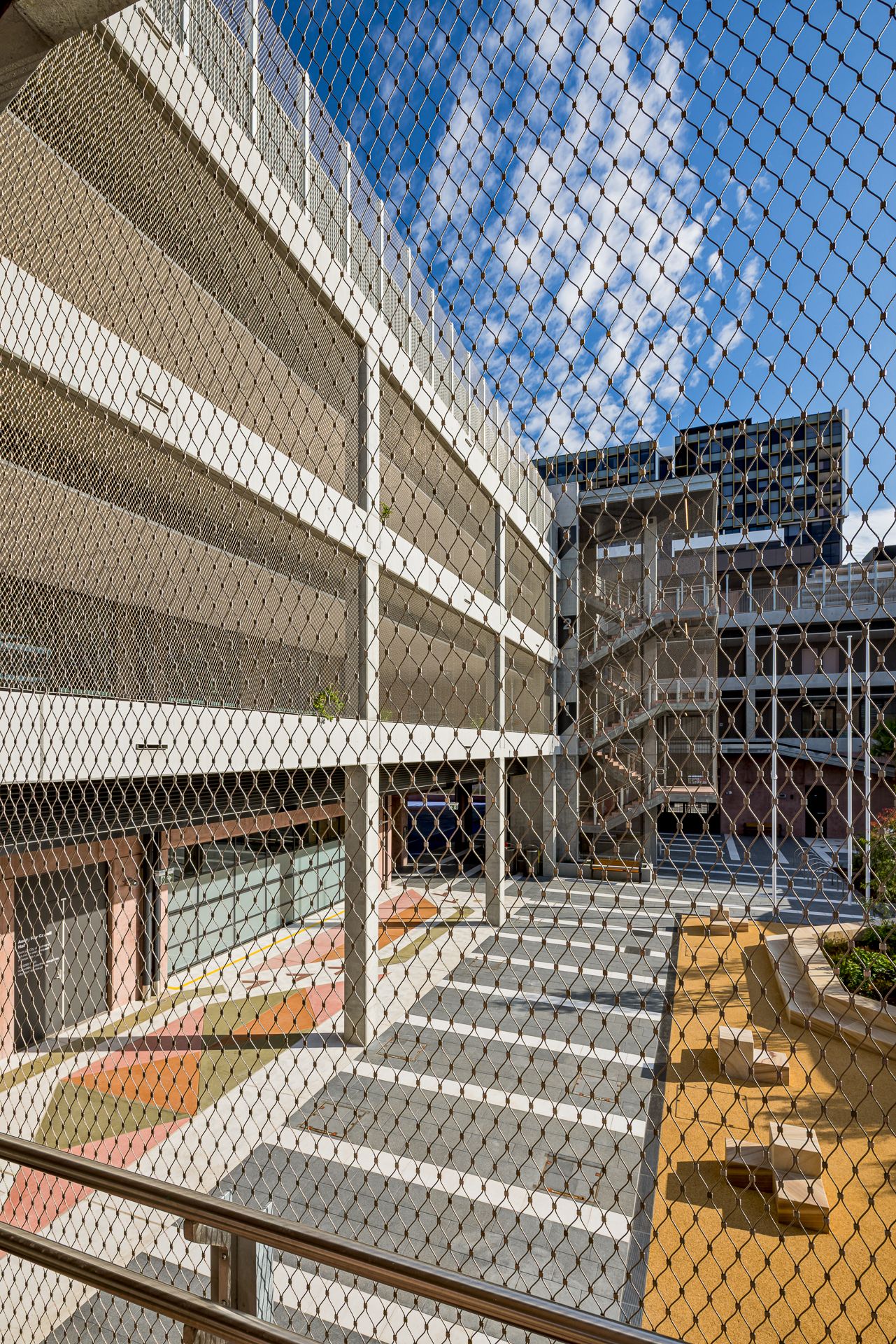 Mesh-enclosed outdoor space beside a multi-level parking structure, with a colorful painted courtyard and modern buildings under a blue sky.