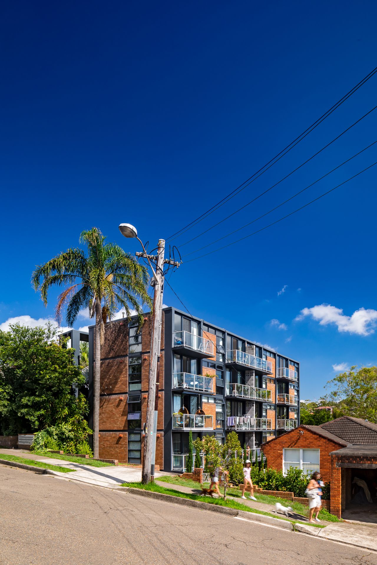 Mid-rise brick apartment building with balconies beside a palm tree and power pole on a sunny street under a deep blue sky.