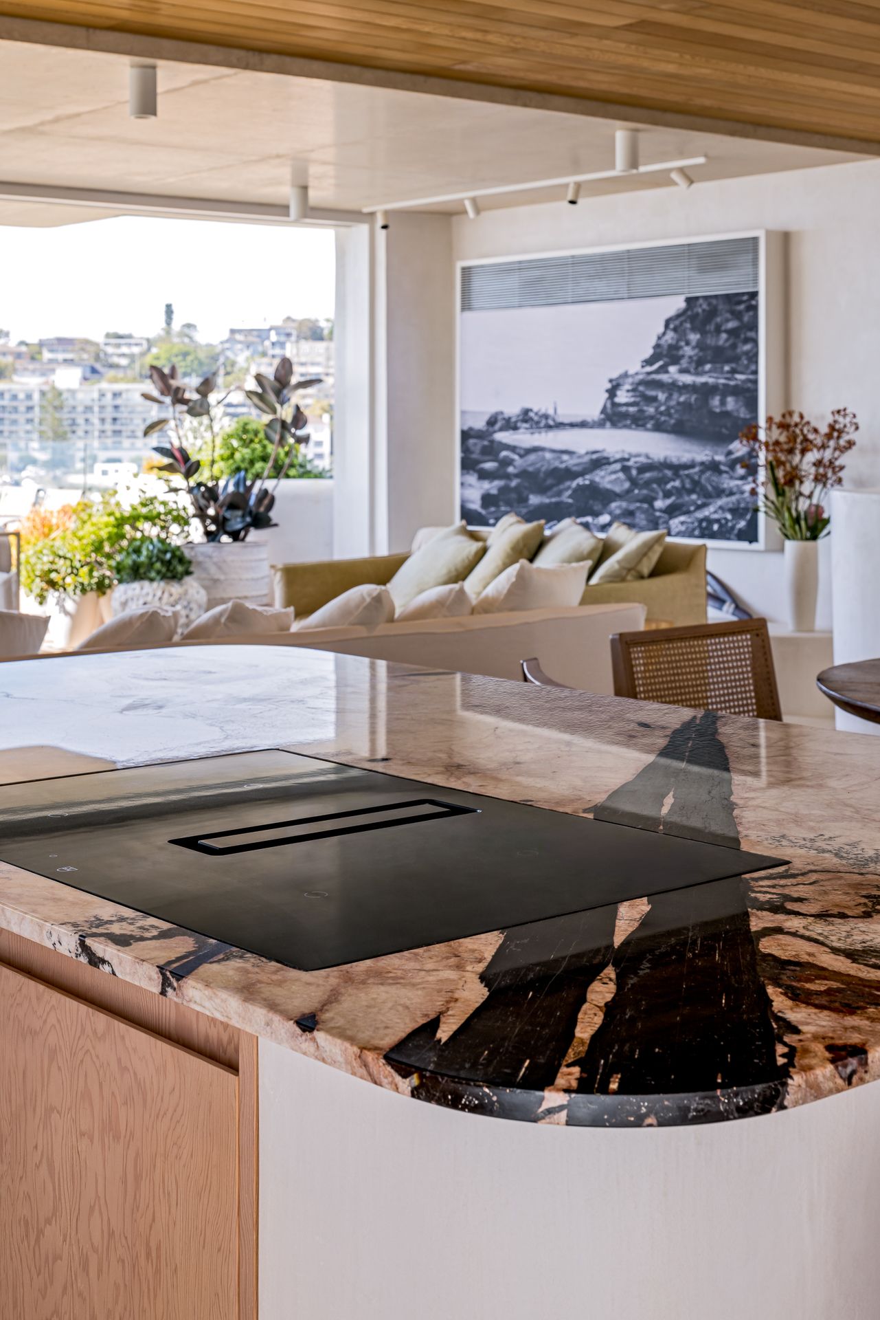 Kitchen island with a veined stone countertop and cooktop, overlooking a lounge, plants and a black and white coastal artwork.