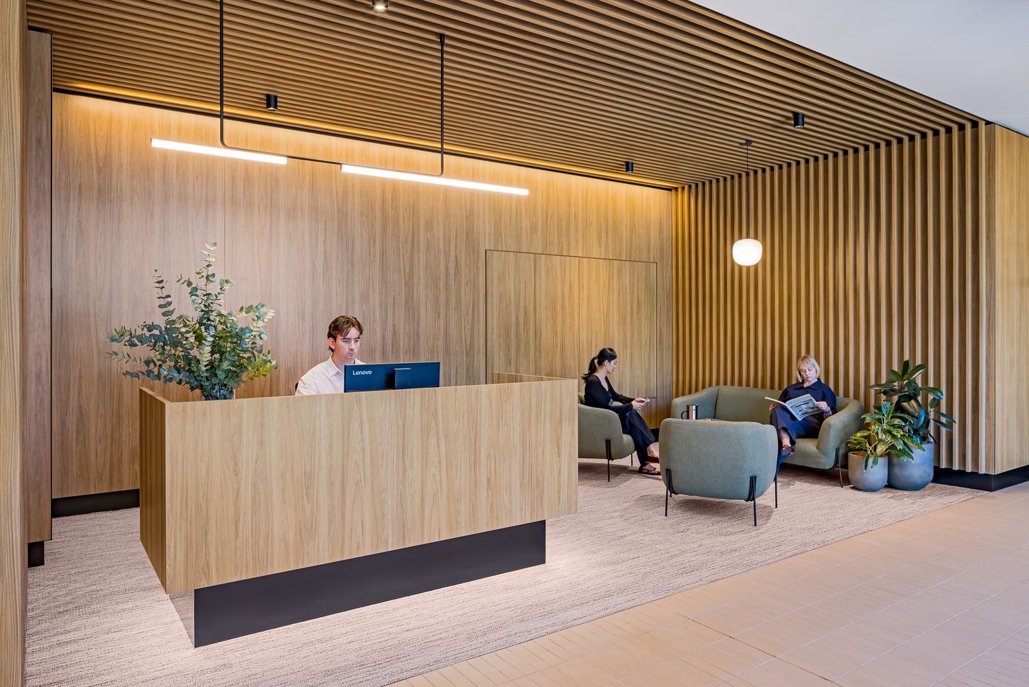 Modern office reception lobby with a wood-paneled desk, receptionist at a computer, pendant lighting, and visitors seated on gray armchairs.