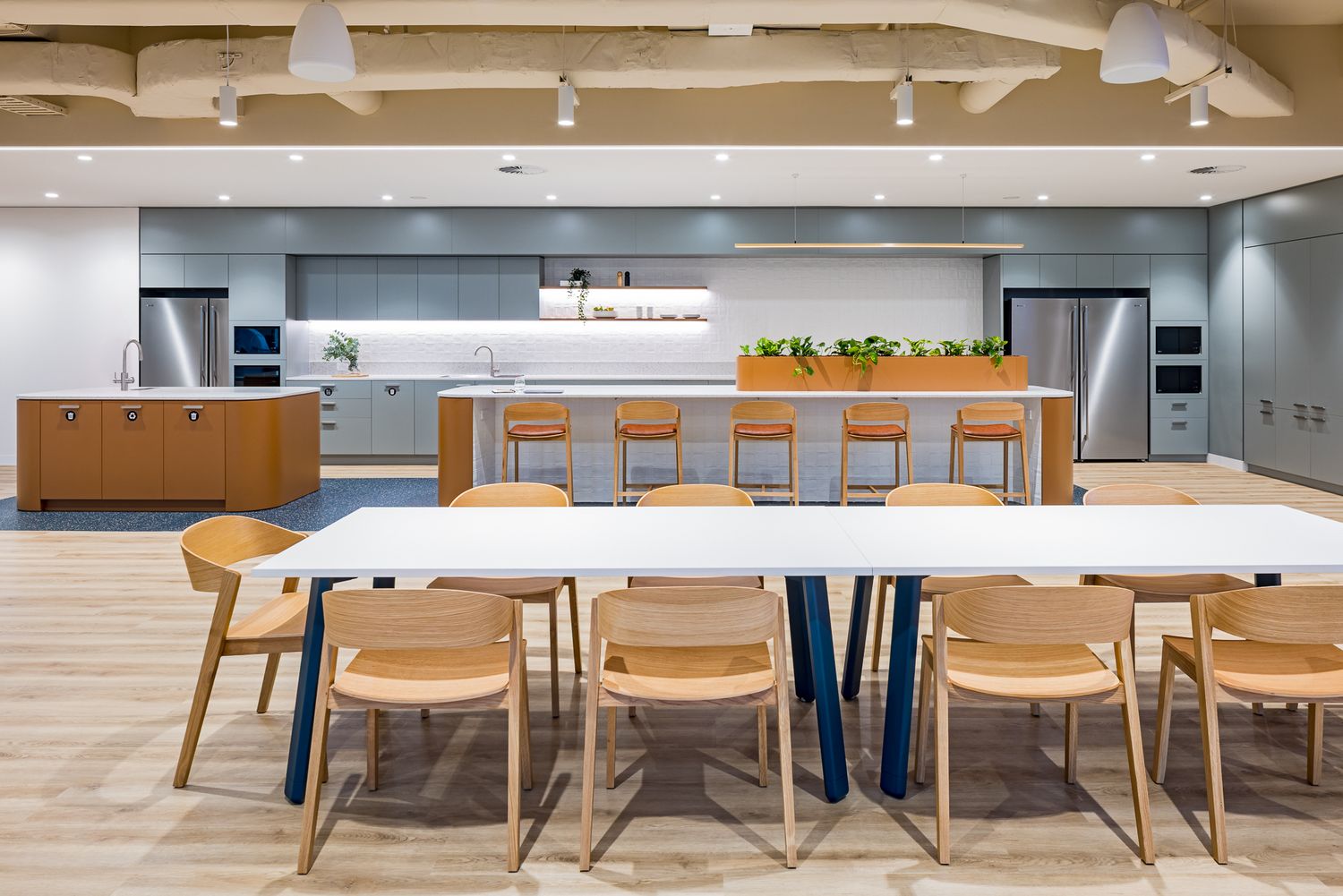 Contemporary office kitchen with a long white communal table, wooden chairs, bar stools, timber cabinetry and stainless steel appliances.