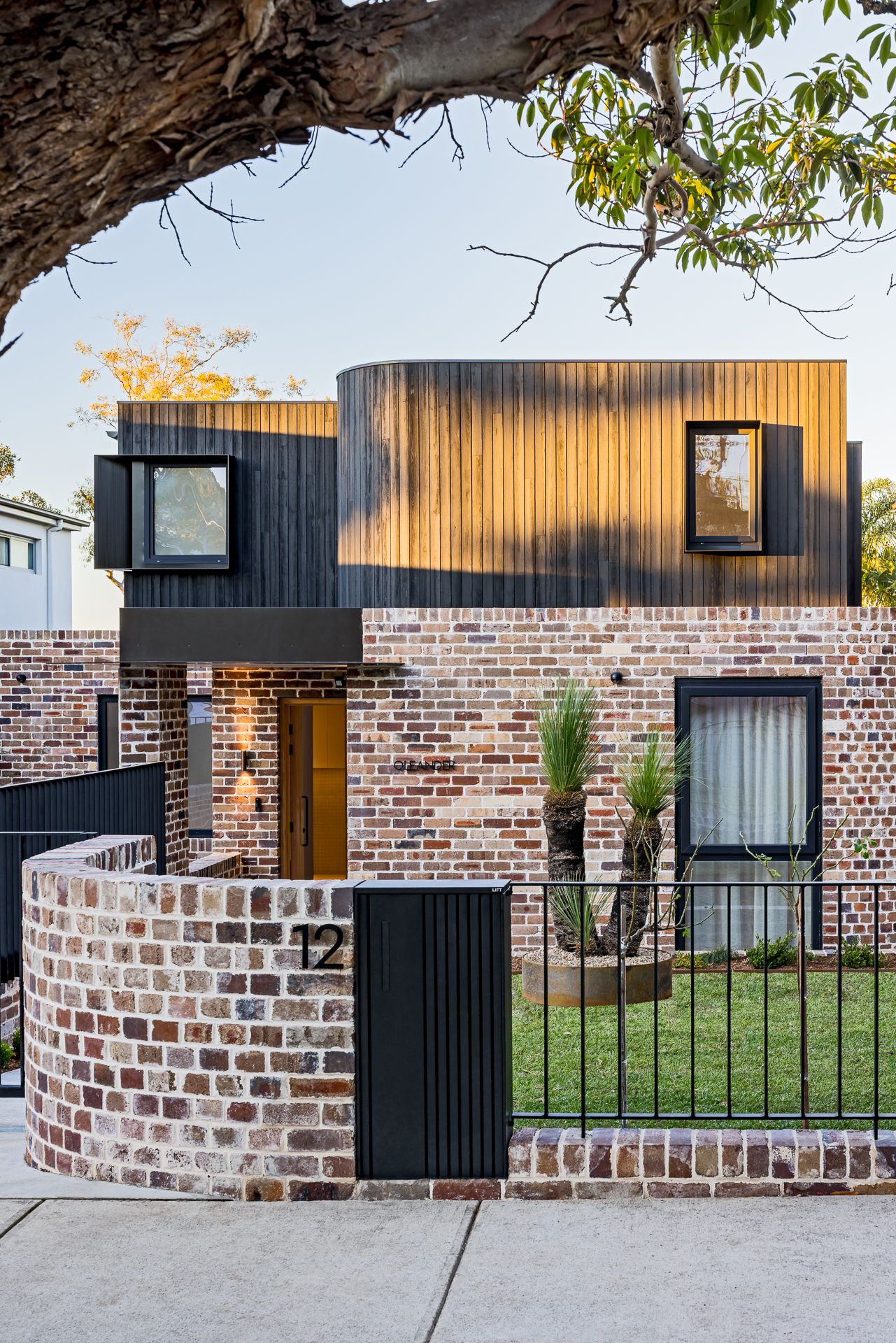 Street view of a modern brick house numbered 12 with a curved brick wall, black timber cladding and a front garden under a gum tree.
