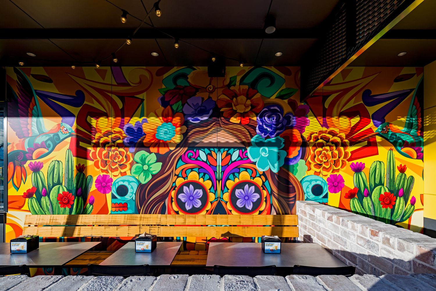 Vibrant floral mural with purple and orange flowers painted above outdoor dining tables at a Mexican restaurant.