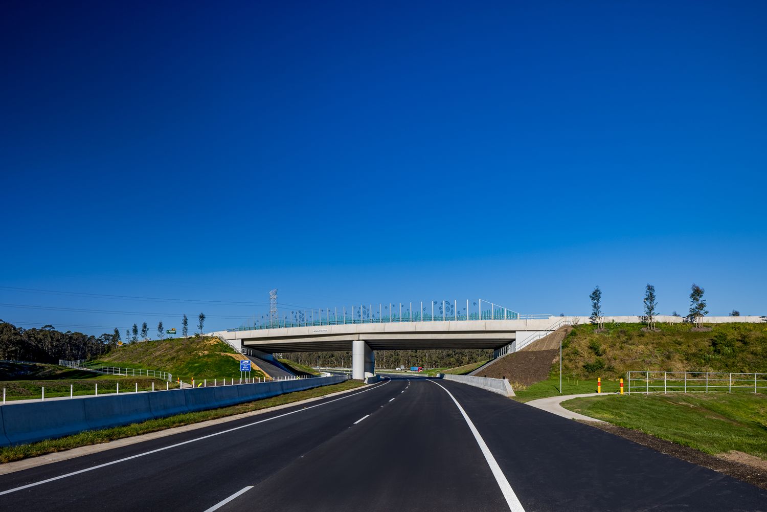 Newly built highway curving under a concrete overpass with noise barriers beneath a clear blue sky.